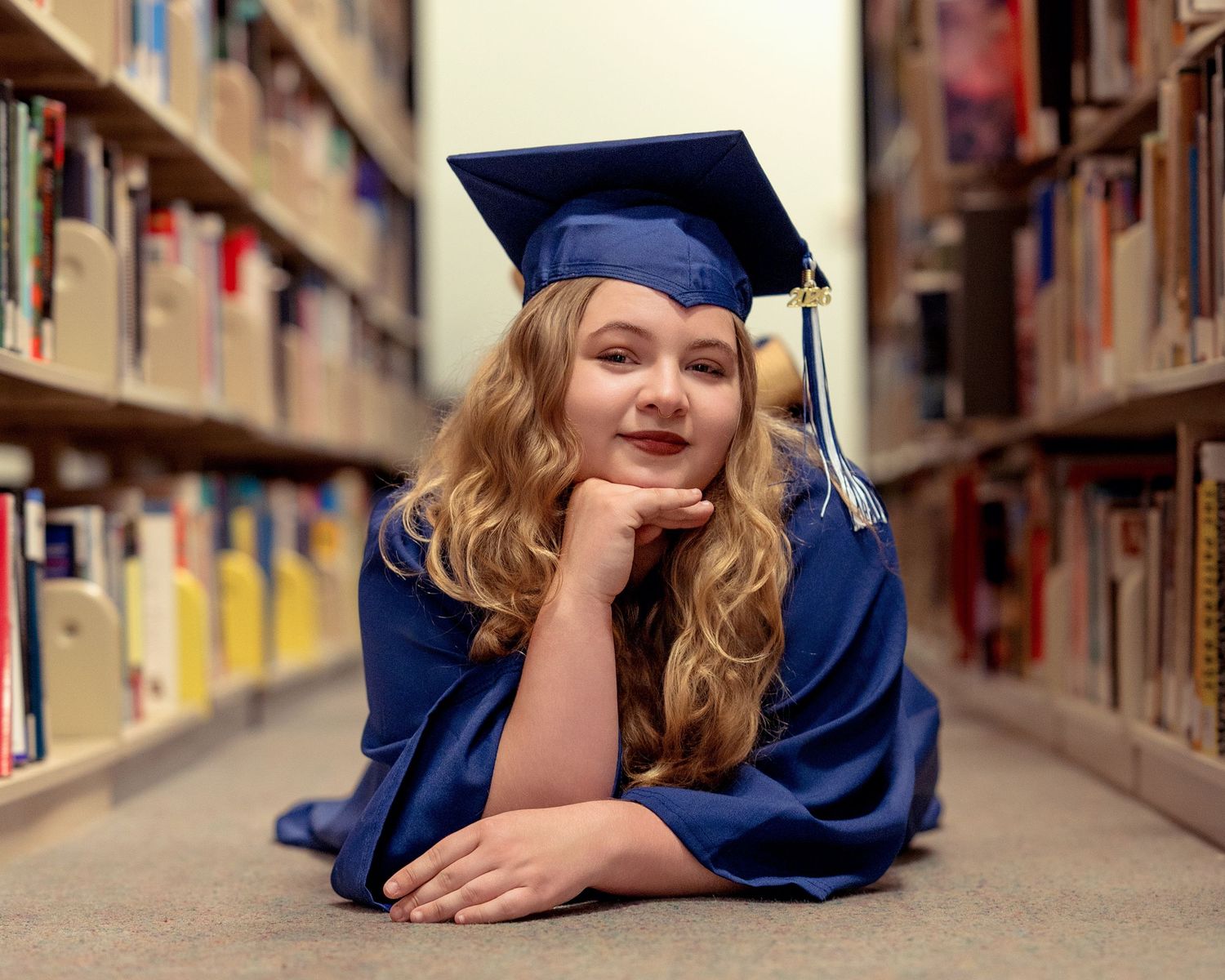 Blonde graduate in blue cap and gown lying on library floor smiling at camera.