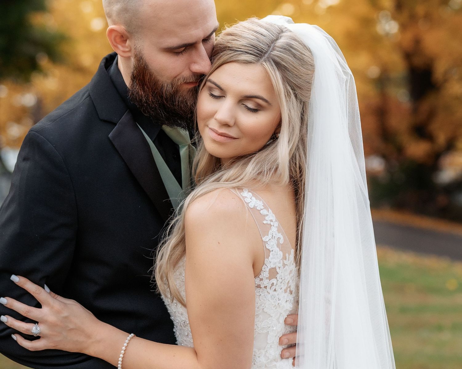Bearded groom in black suit kisses bride in lace gown and veil among autumn foliage outdoors.