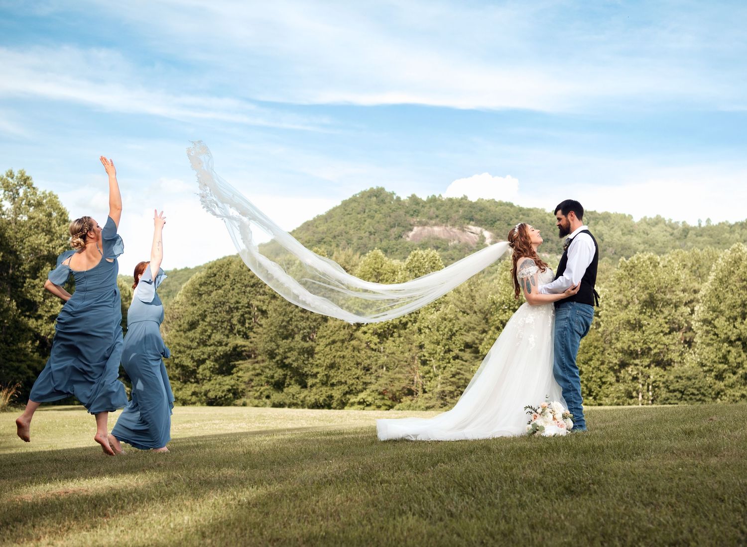 Bride and groom kissing while bridesmaids jump joyfully as veil flows in mountain meadow setting.