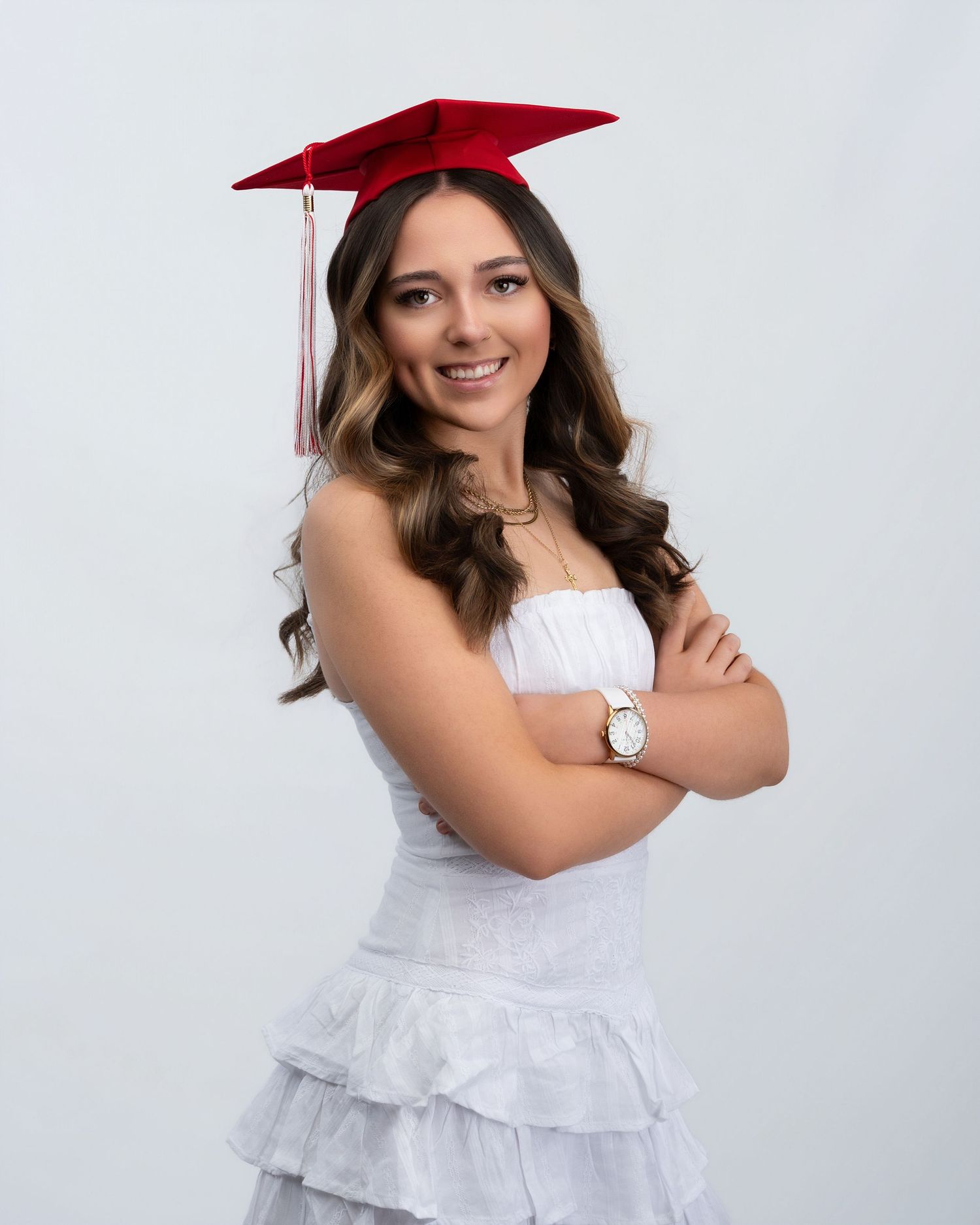 Smiling young woman in red graduation cap and white dress with arms crossed for senior portrait.