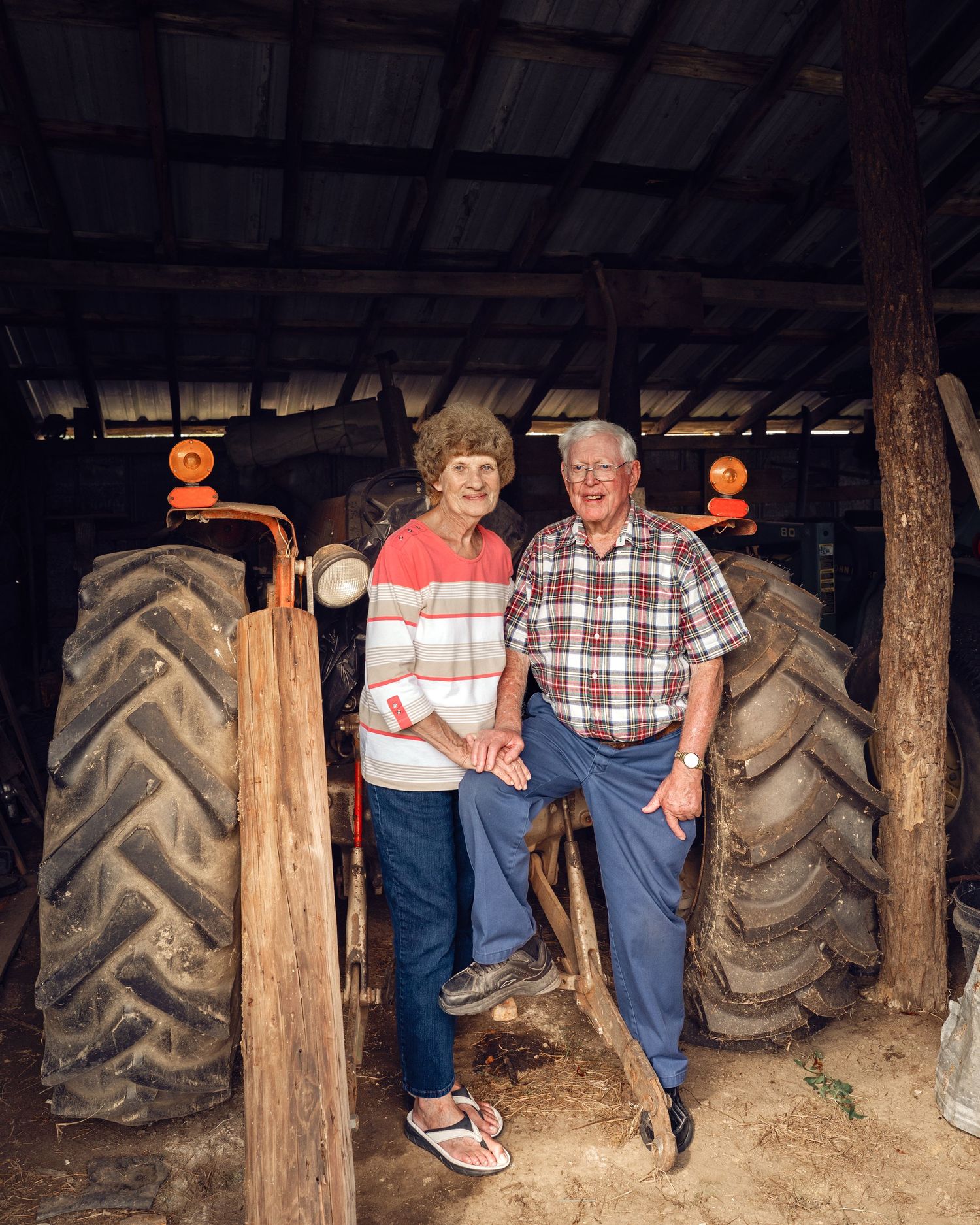 Senior couple smiling together beside a vintage red tractor inside a rustic wooden barn.