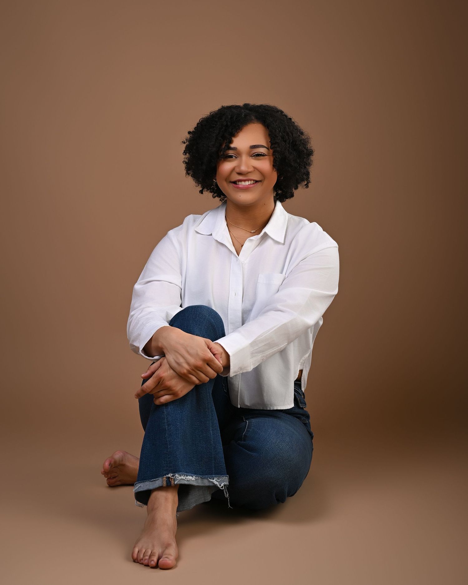 A smiling woman with curly hair sits cross-legged barefoot in white shirt and jeans on tan backdrop.