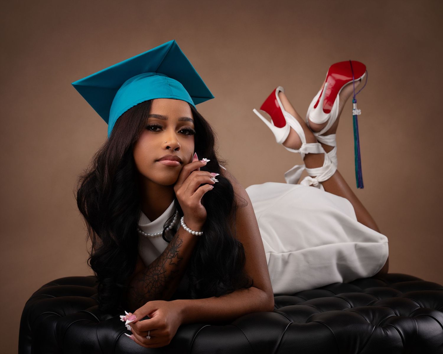 Graduate in teal cap poses on black ottoman in white dress with red-soled heels in studio photo.