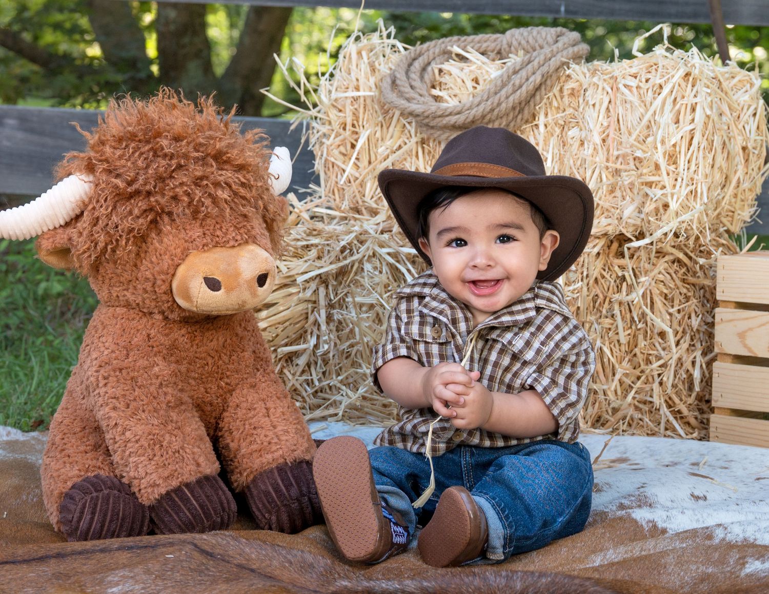 Smiling baby boy in cowboy hat poses with highland cow plush toy beside hay bales outdoors.