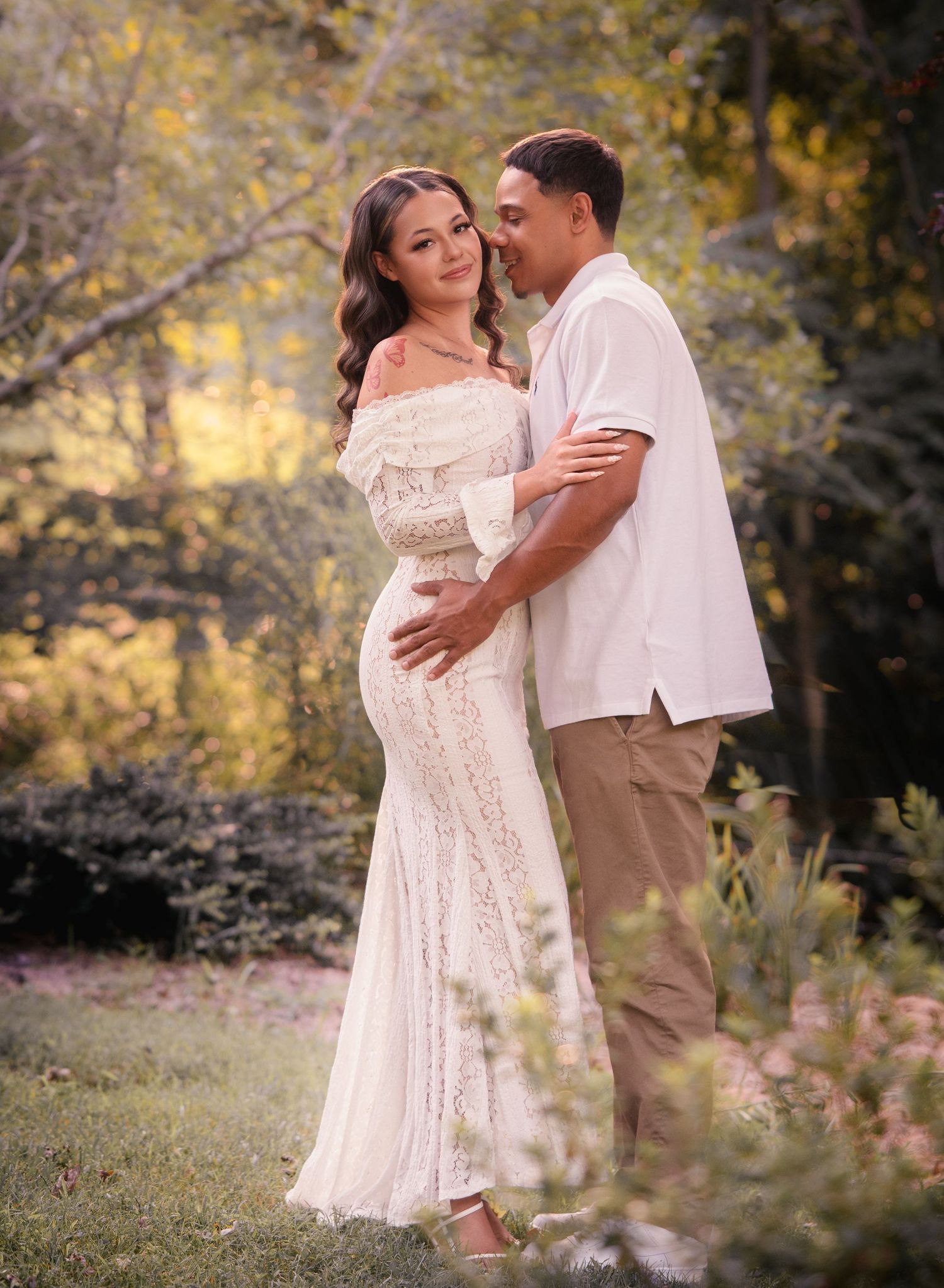 Maternity photo of couple in garden, woman in white lace gown with partner in neutral outfit.