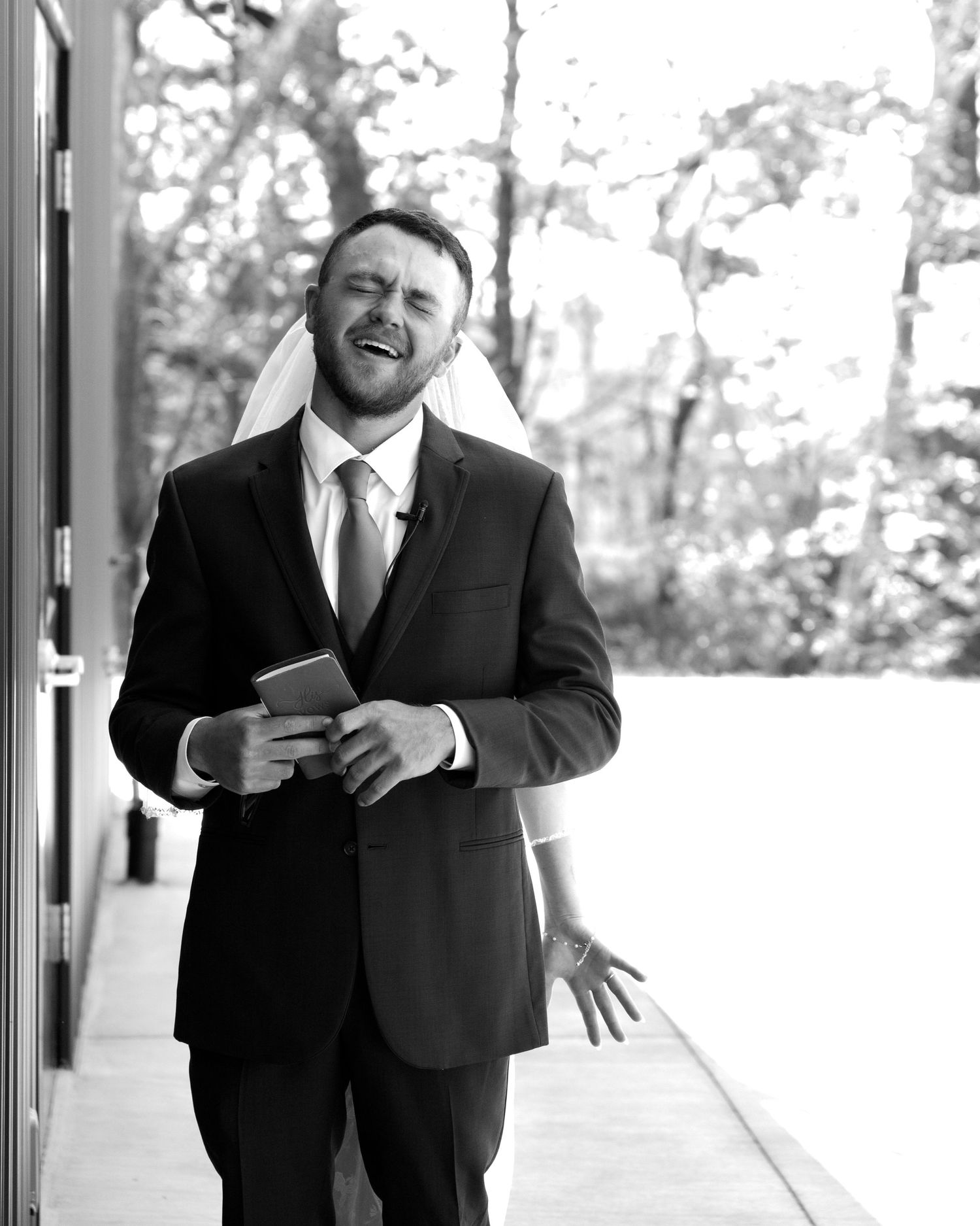 Joyful groom in black suit laughing while holding a book outdoors on his wedding day in black and white.