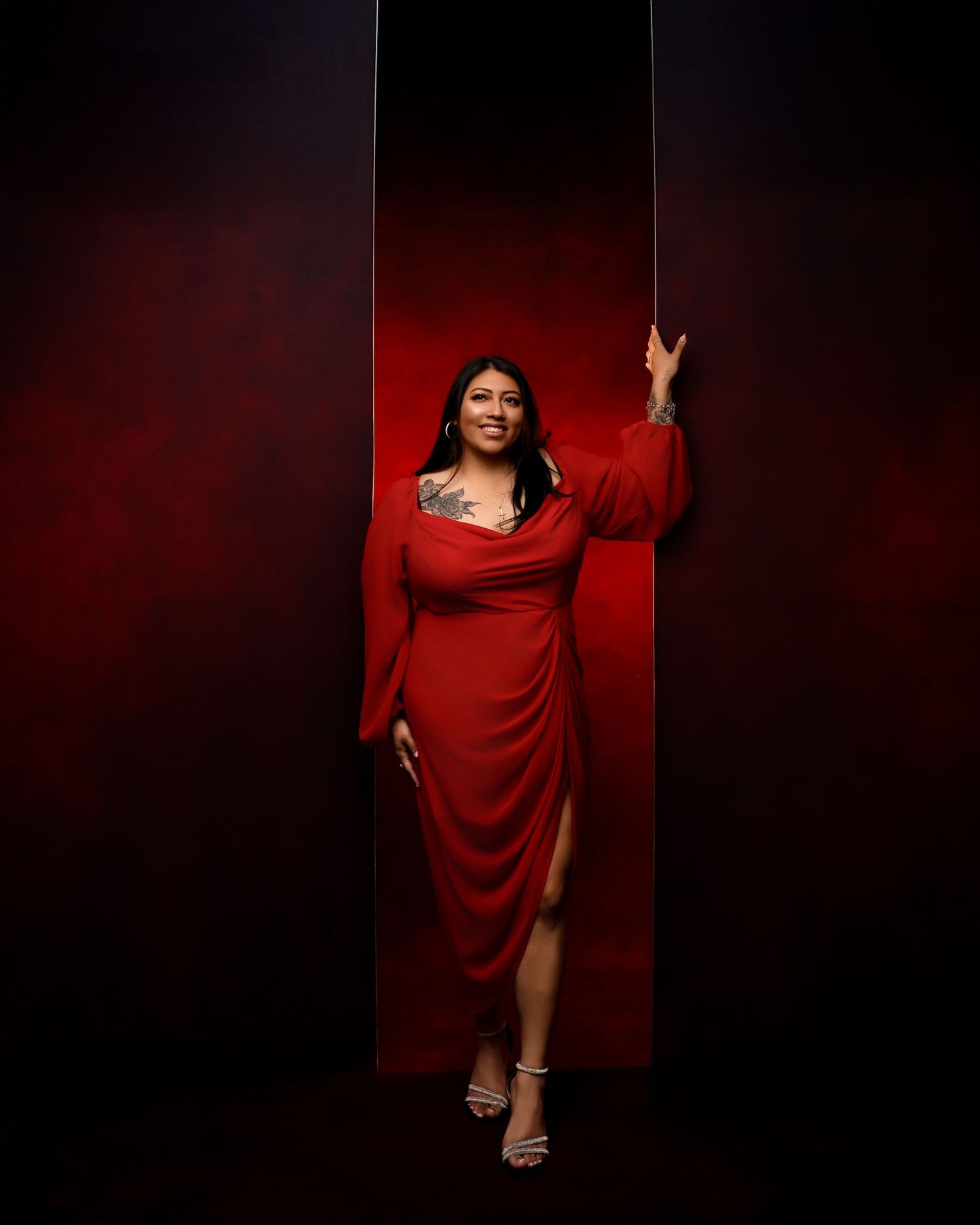 Woman in a flowing red wrap dress with a high slit poses against a dramatic red and black backdrop, one hand raised, wearing strappy heels.
