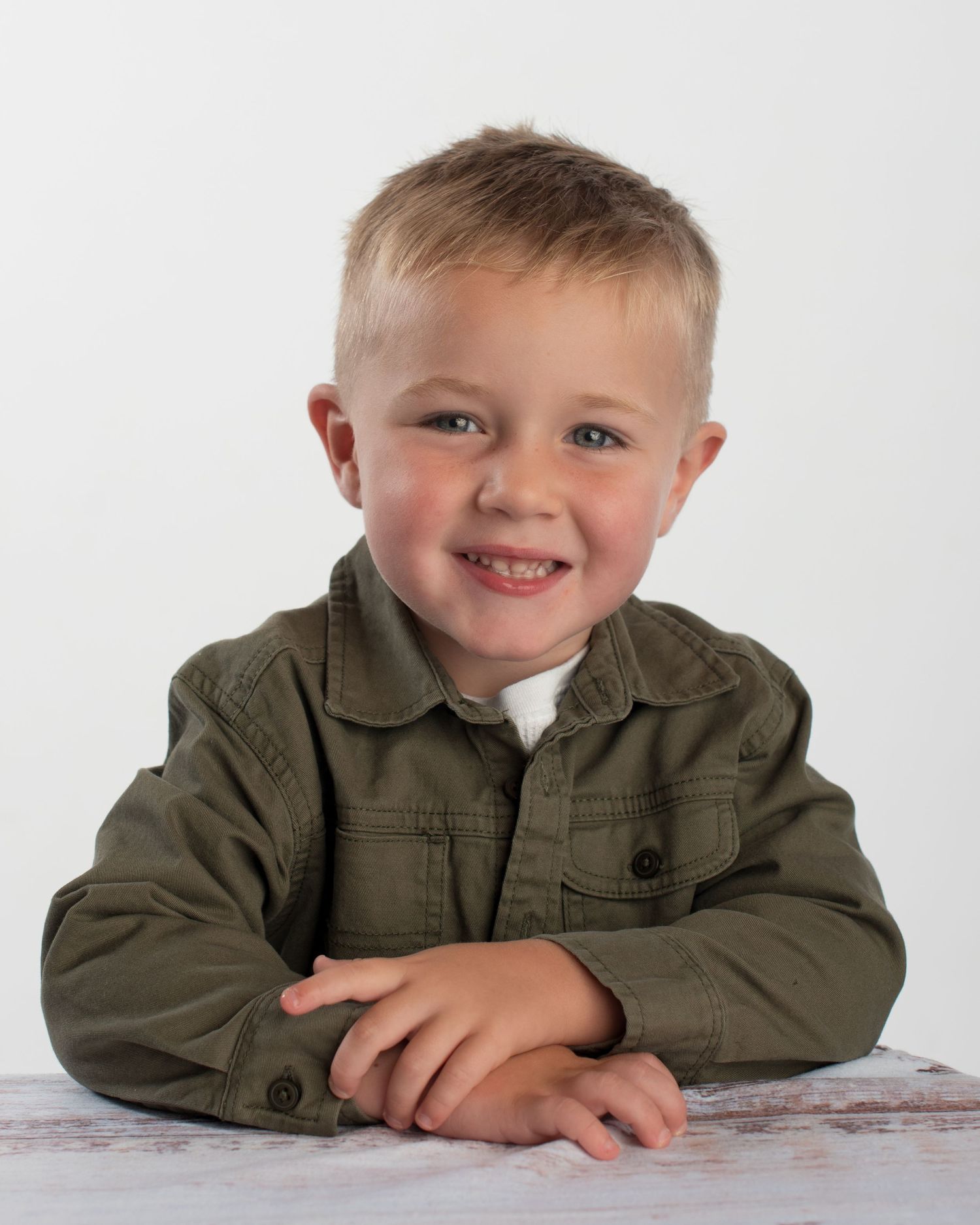 Adorable blonde toddler boy in olive green button-up shirt smiling brightly in professional studio portrait.