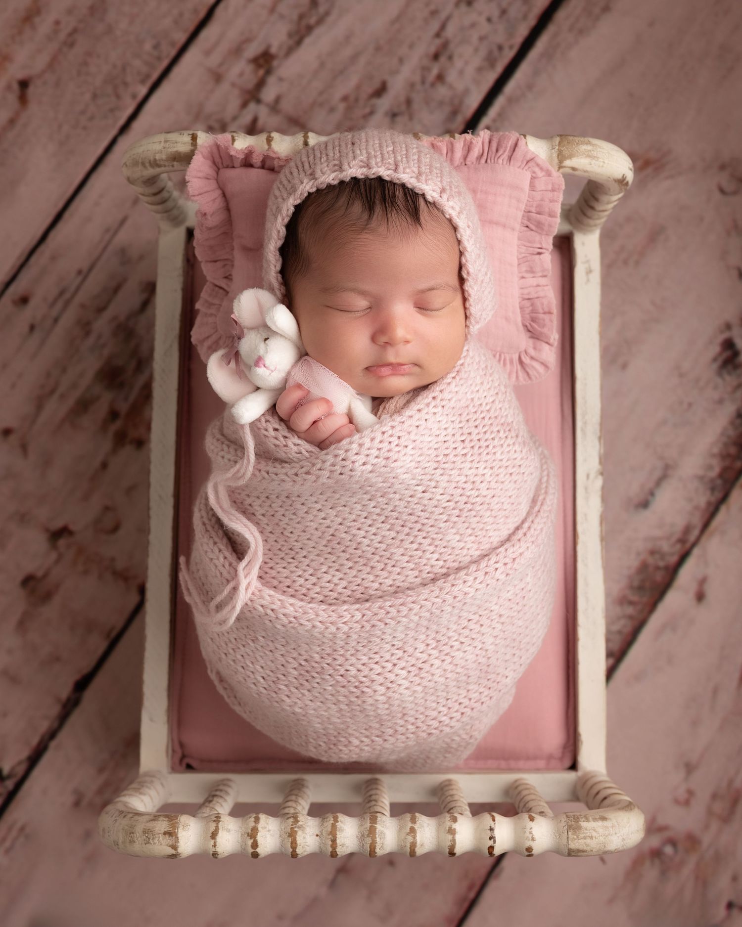 Sleeping newborn baby in pink knit swaddle and bonnet lying in a small decorative wooden bed.
