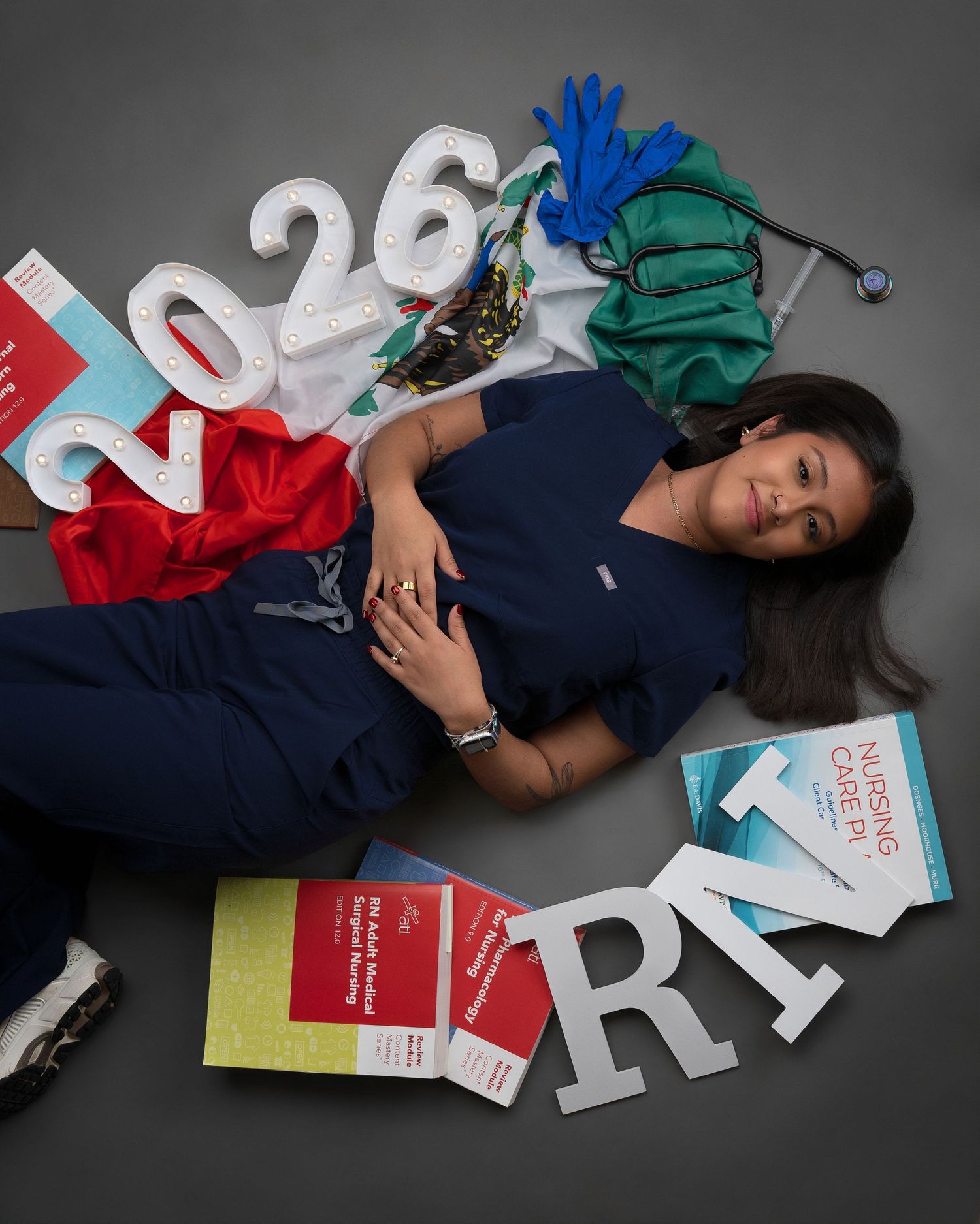 Nursing student in scrubs lies surrounded by textbooks, RN letters, and 2026 graduation props.