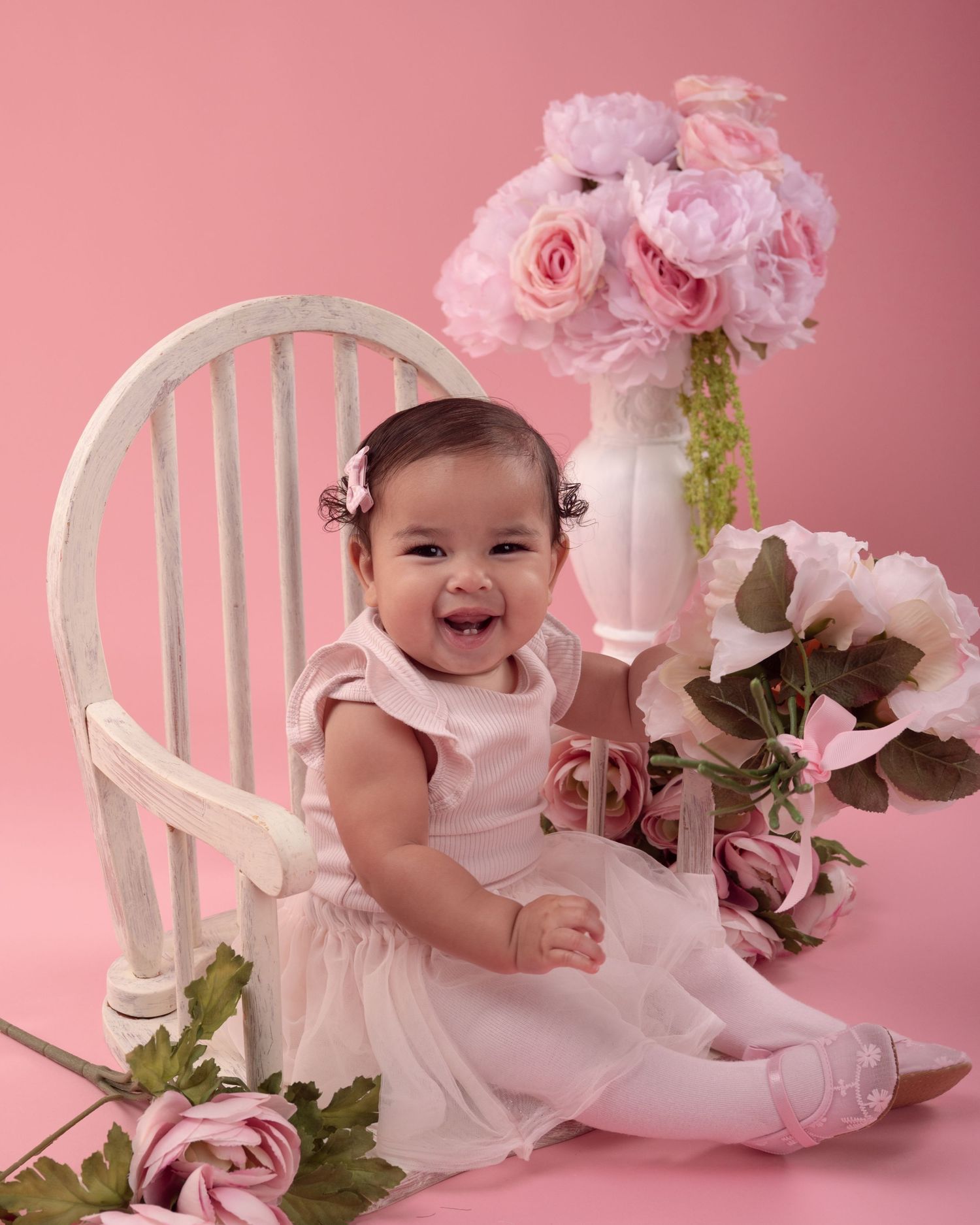 Smiling baby girl in pink dress sitting with roses and flowers in a pink studio portrait.