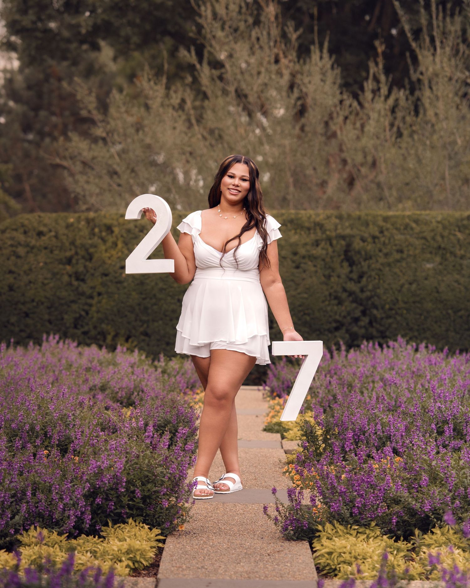 Young woman in white dress posing with number 27 signs in lavender garden for graduation photo.