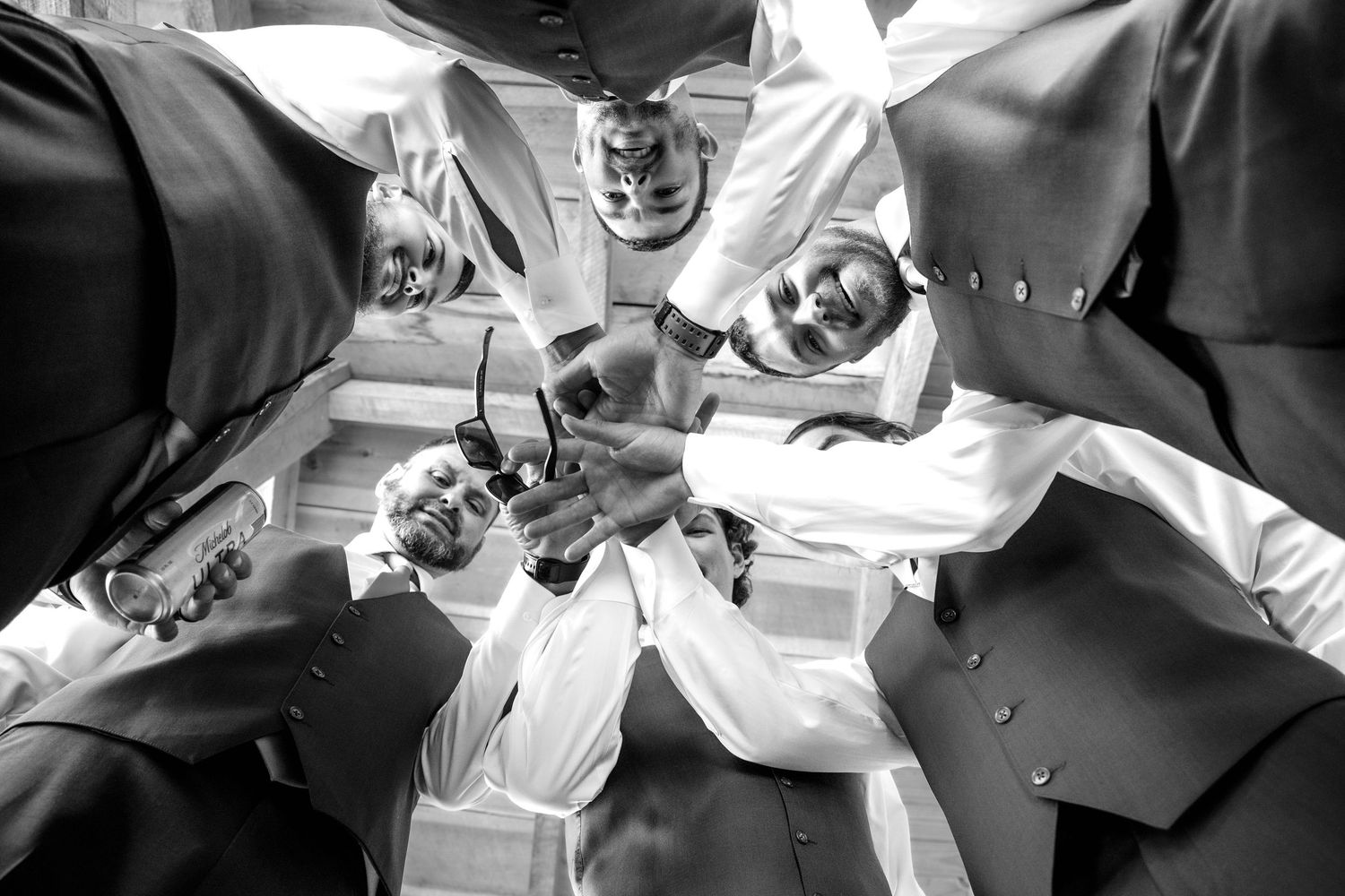 Black and white low angle shot of groomsmen in a huddle toasting with sunglasses on wedding day.