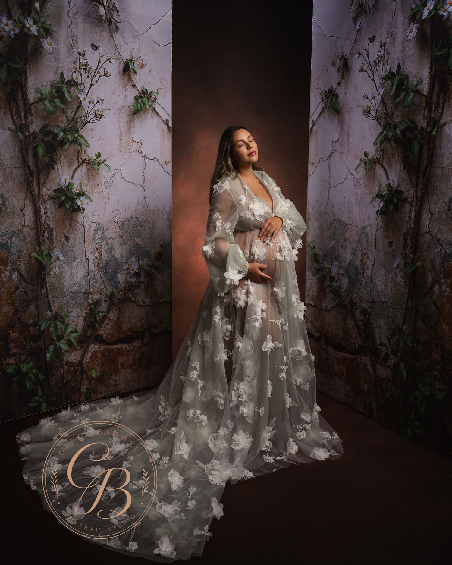 Pregnant woman in sheer floral sage green maternity gown posing in elegant studio with vine-covered walls.