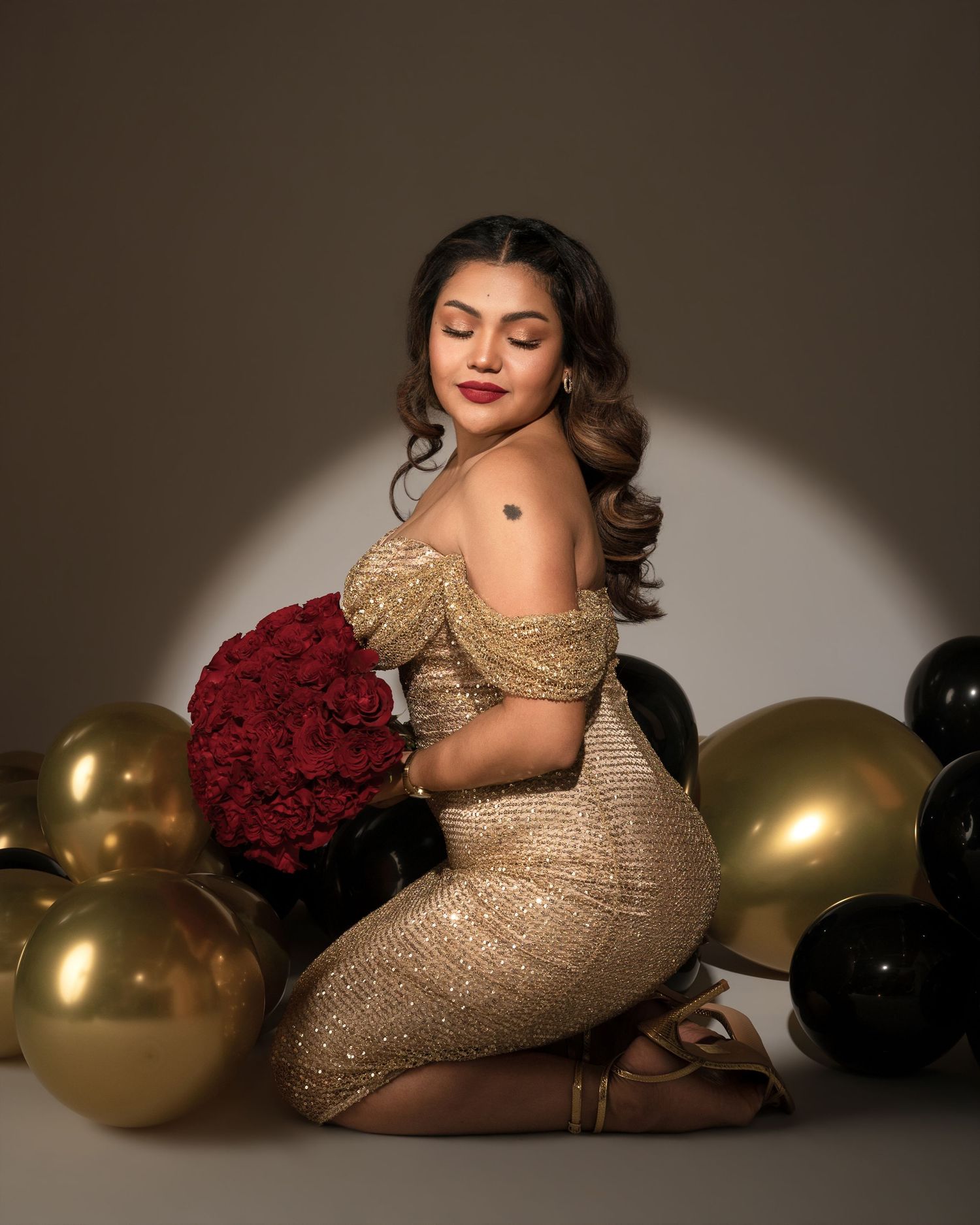 Woman in a gold sequined off-shoulder dress kneels holding a red rose bouquet, surrounded by gold and black balloons in a spotlit studio.