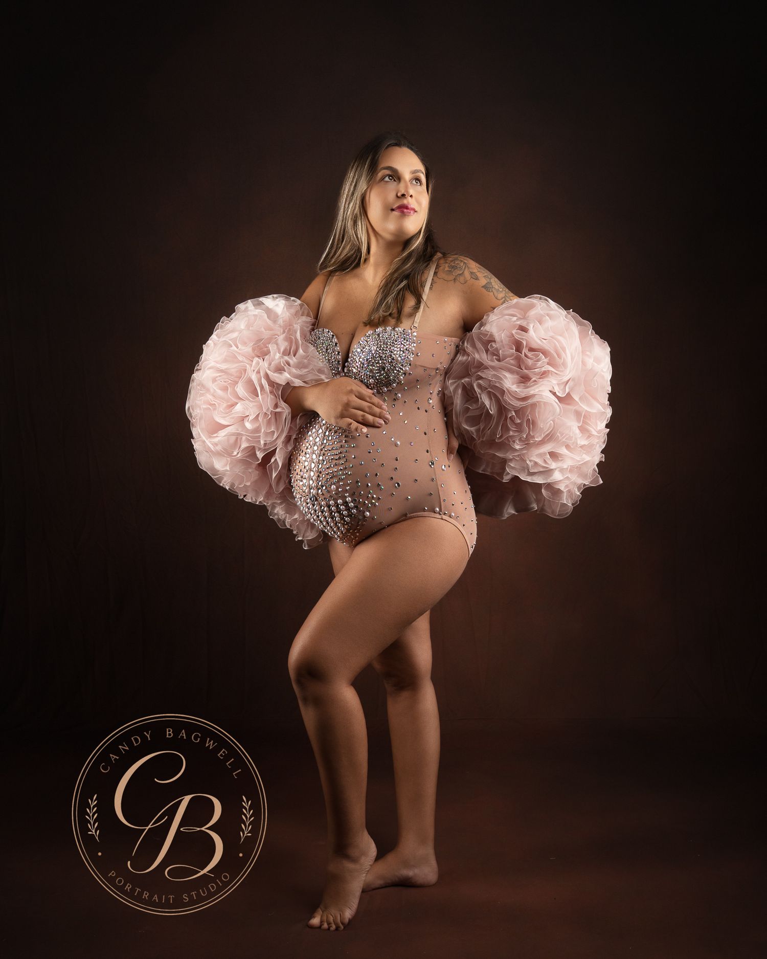 Pregnant woman poses in a pink ruffled maternity bodysuit with rhinestone details for a professional studio portrait.