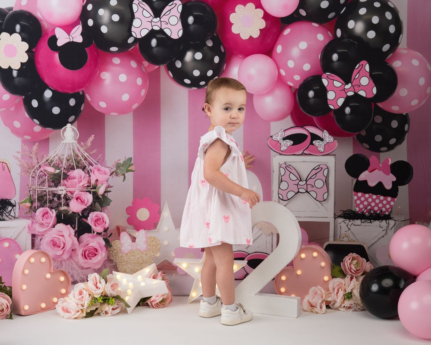 A baby girl in a white dress stands before a pink and black Minnie Mouse balloon arch birthday backdrop.
