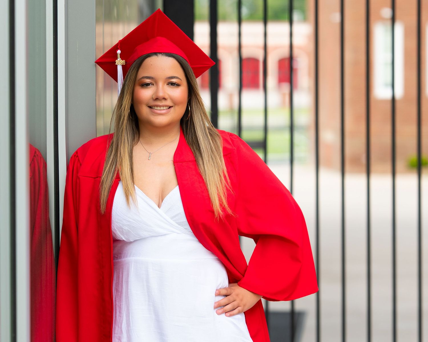 Pregnant graduate smiles in red cap and gown with white dress, posing at school entrance doors.