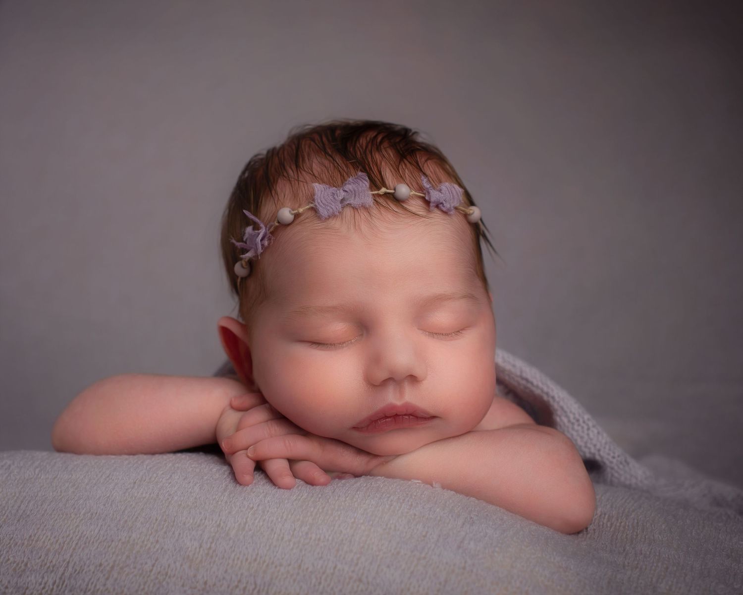 Peaceful sleeping newborn baby girl with lavender bow headband resting chin on hands.