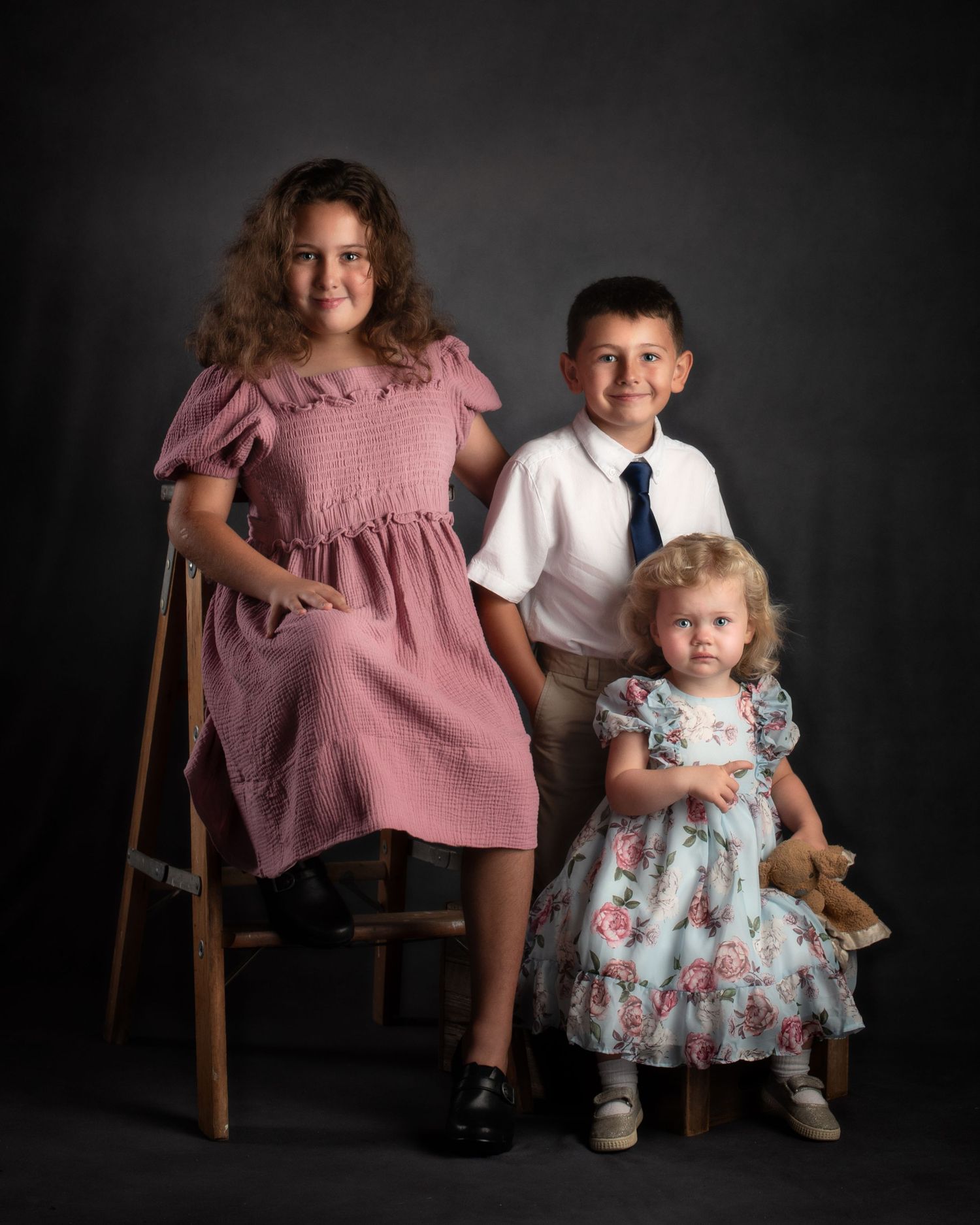 Three children in formal attire pose for classic studio portrait against dark gray background.