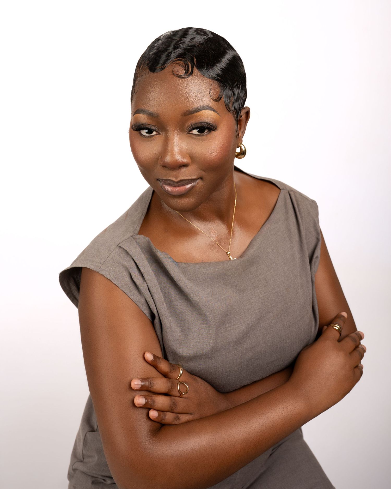 Professional woman in gray sleeveless top with crossed arms, wearing gold jewelry, posing confidently against white background.