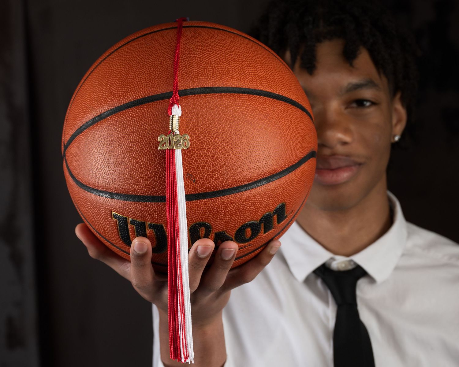Young man in white shirt and tie holds Wilson basketball with red graduation tassel in senior portrait.
