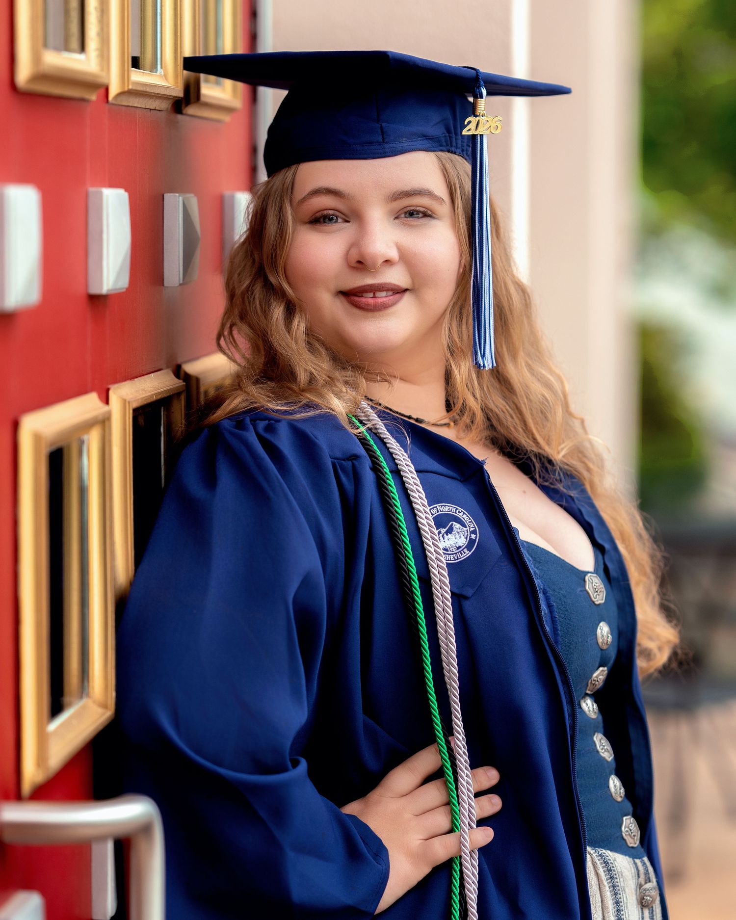 Smiling female graduate in blue cap and gown with honor cords posing against a red wall outdoors.