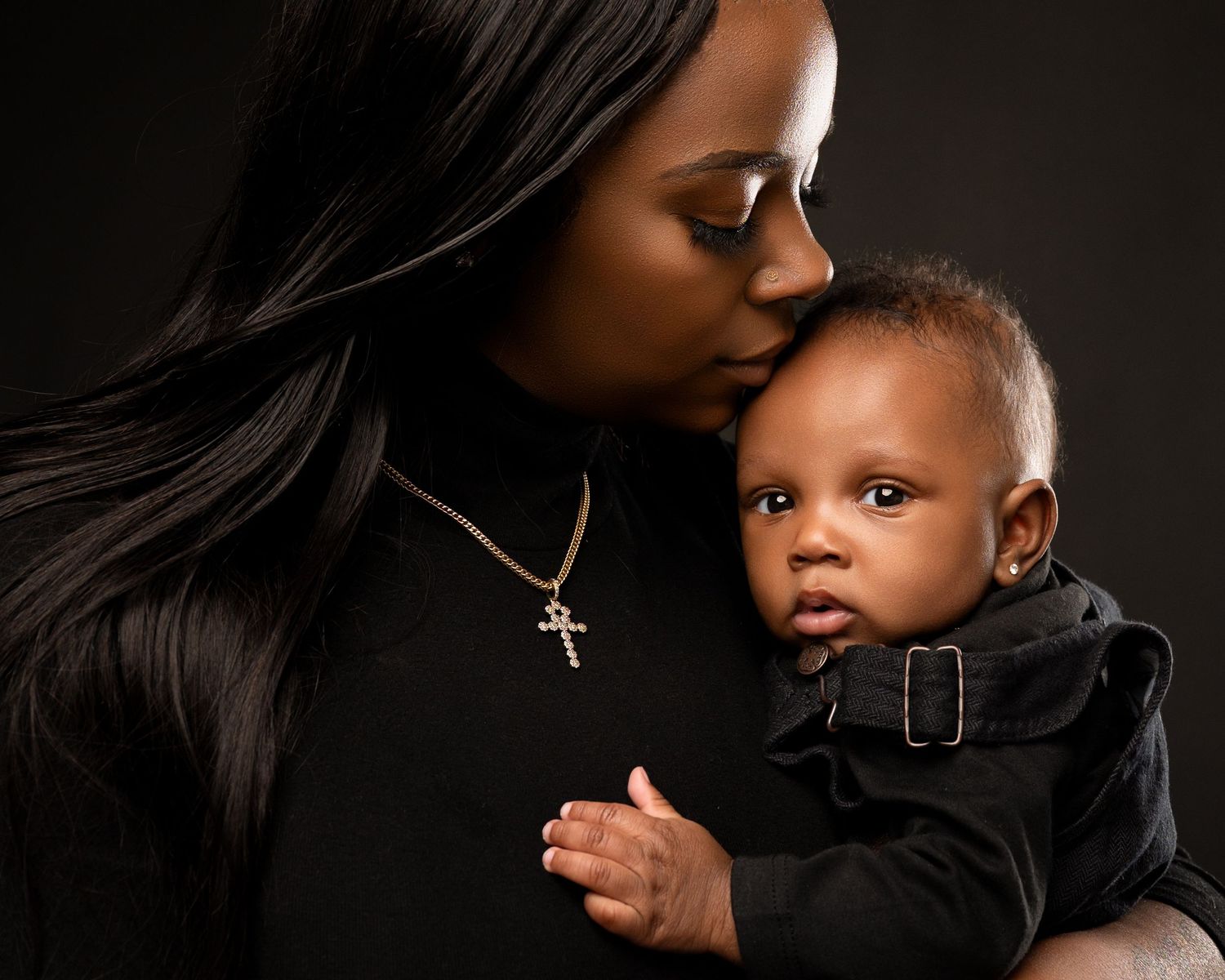 A mother kisses her baby's head while holding him close in a dark studio portrait.