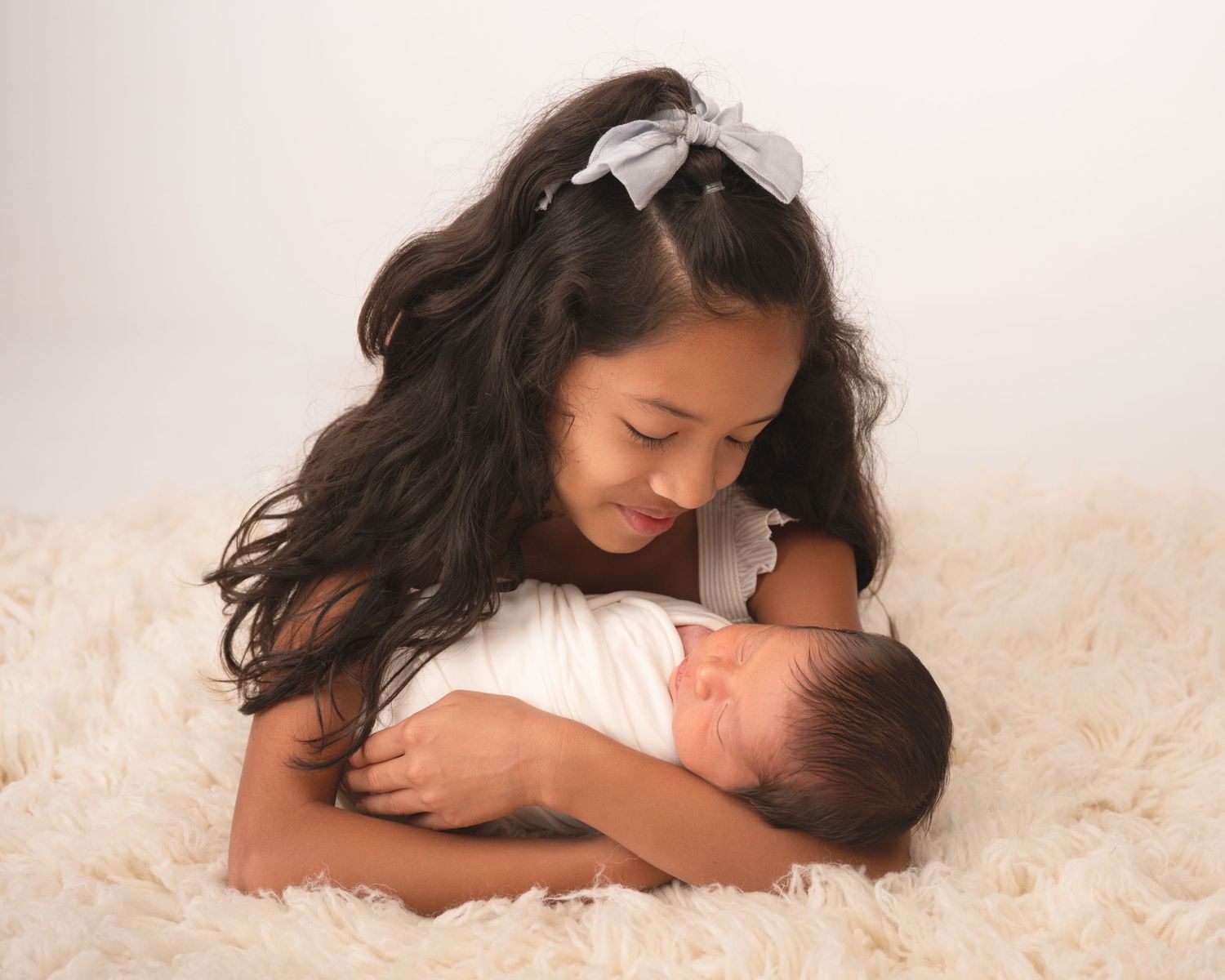 A young girl with a bow headband tenderly holds a sleeping newborn baby on a fluffy cream rug.