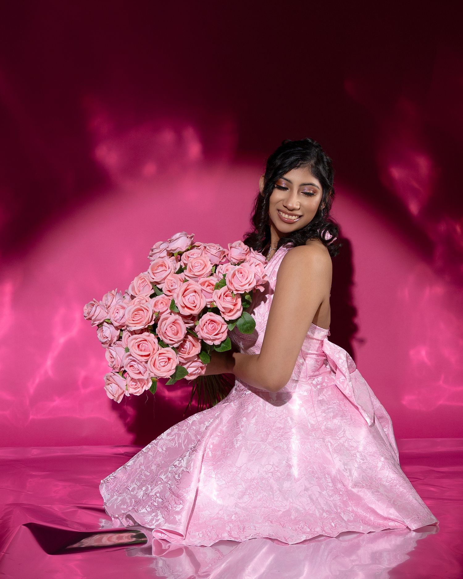 Young woman in pink ball gown holding large bouquet of pink roses on pink studio background.
