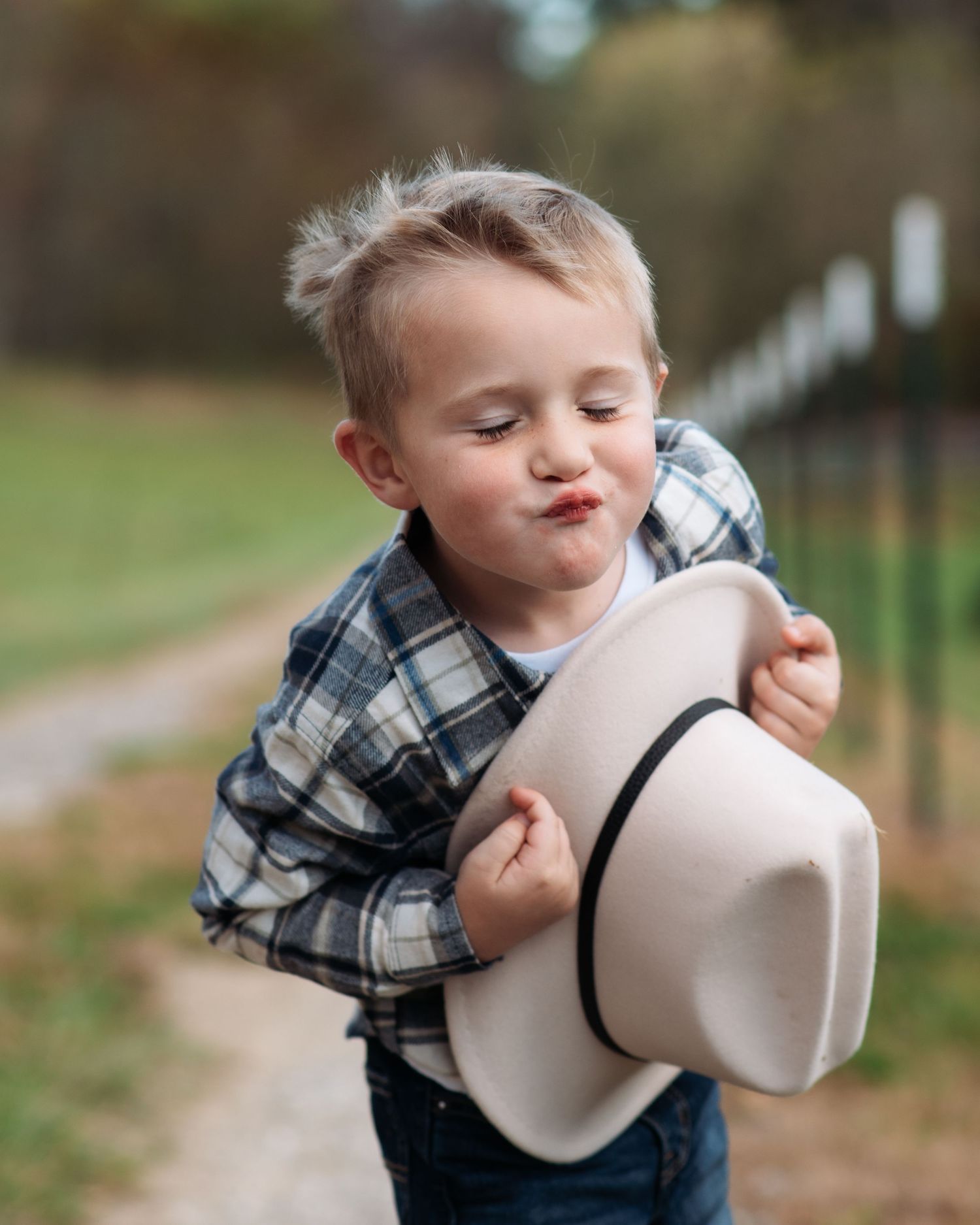 Cute toddler boy in plaid shirt making funny face while holding a cowboy hat outdoors.