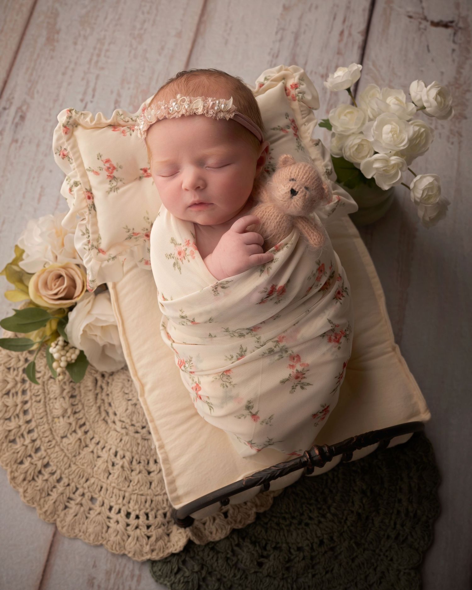Sleeping newborn swaddled in floral wrap on miniature bed with teddy bear roses and white flowers.