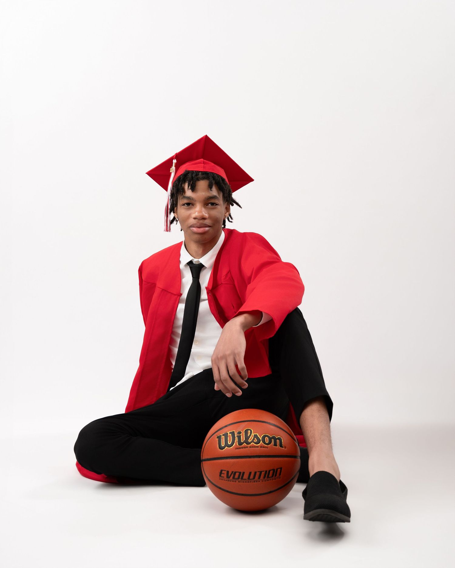 Male graduate in red cap and gown sitting cross-legged with Wilson basketball in studio.