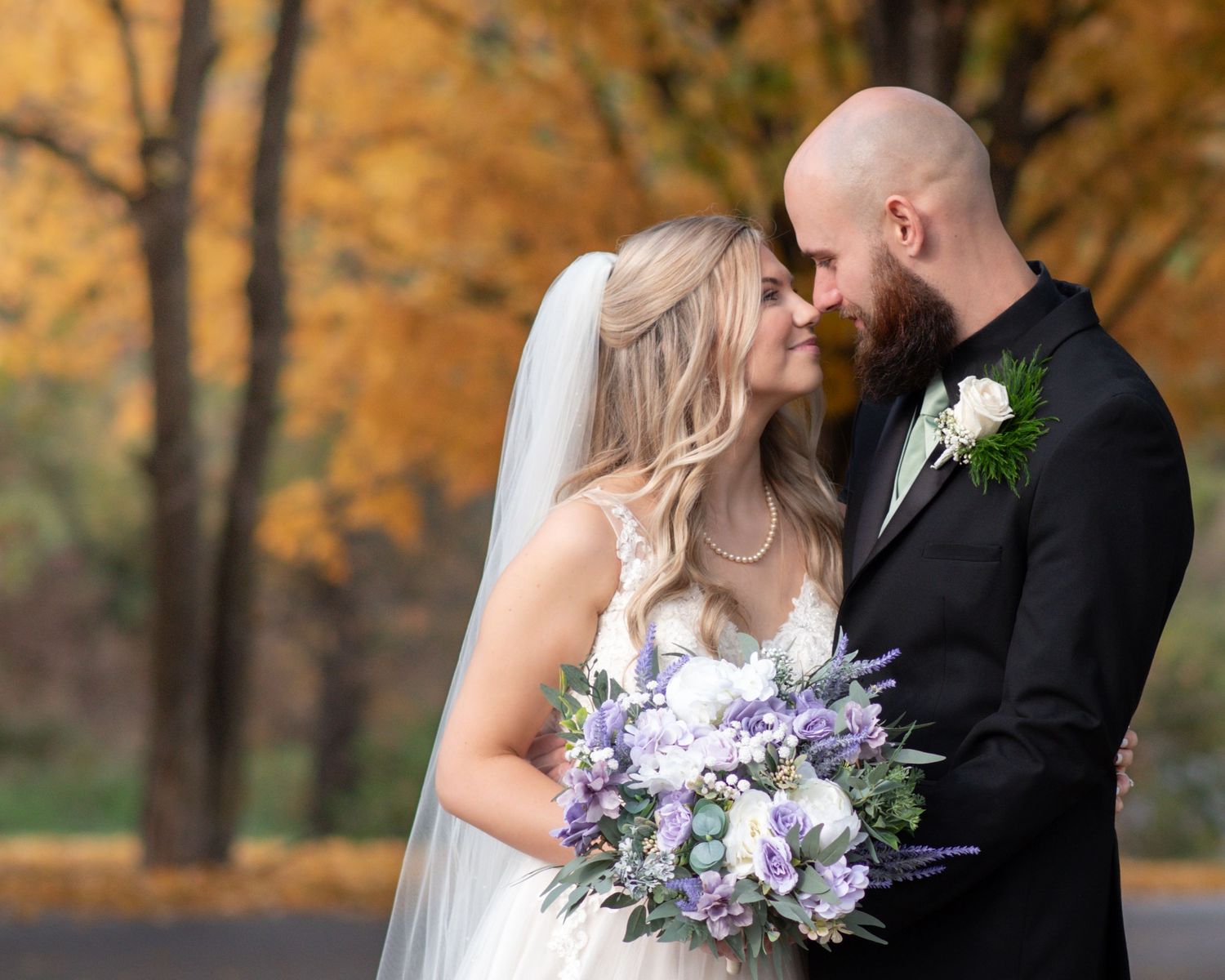 Bride and groom share a tender kiss surrounded by vibrant golden autumn foliage holding purple bouquet.