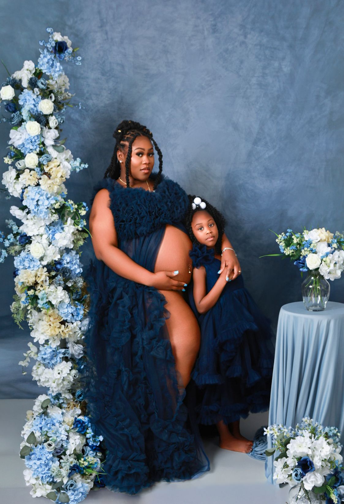 Pregnant woman in navy blue tulle gown poses with young girl in matching dress among blue floral decor.