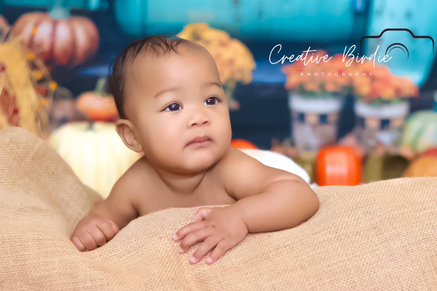 Curious baby lies on burlap surface with festive fall pumpkin backdrop in professional photo session.