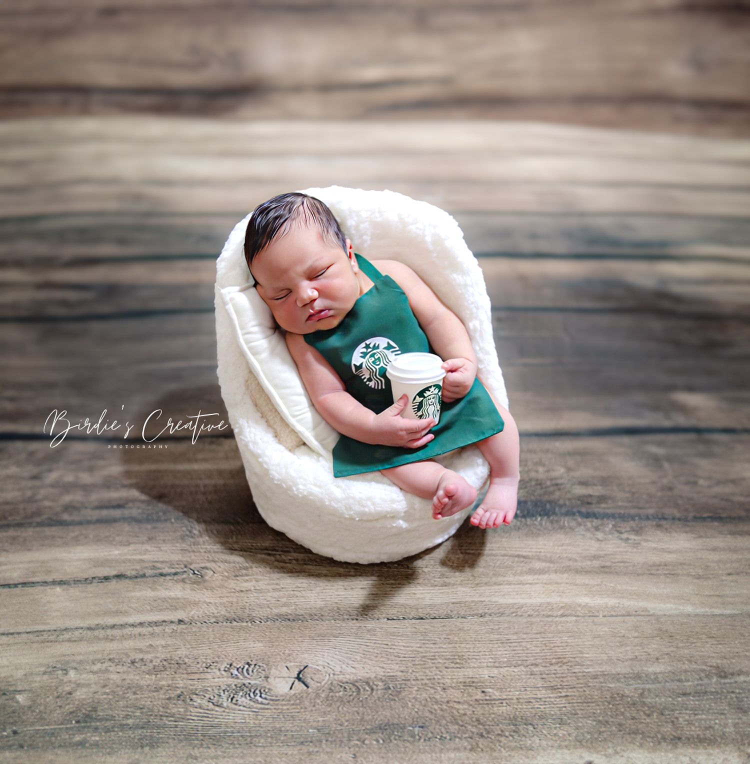 Sleeping newborn baby in a green Starbucks-themed outfit holding a mini cup in a bowl.