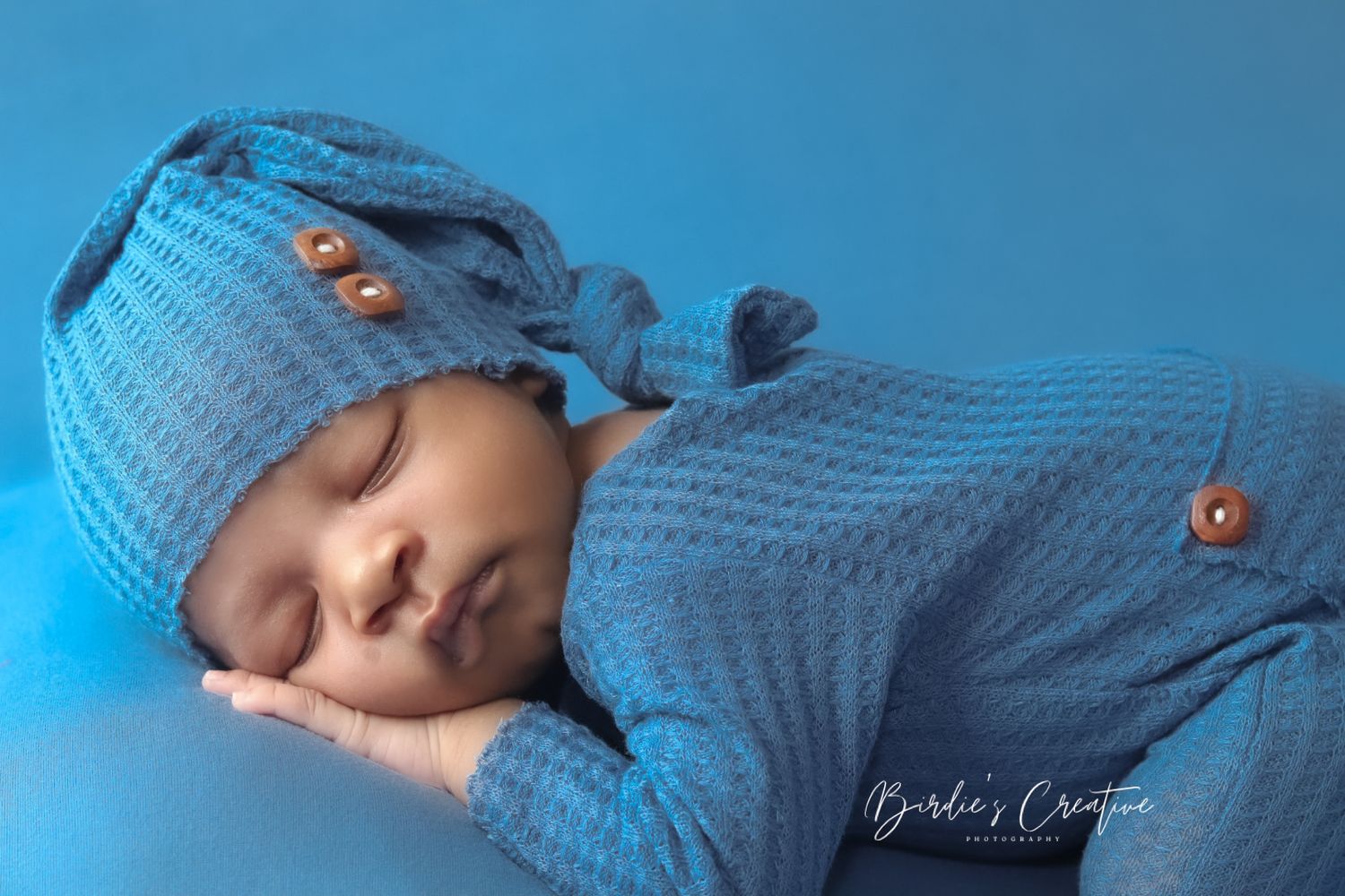 Newborn baby sleeps soundly in blue knit romper and matching hat in professional photography studio.