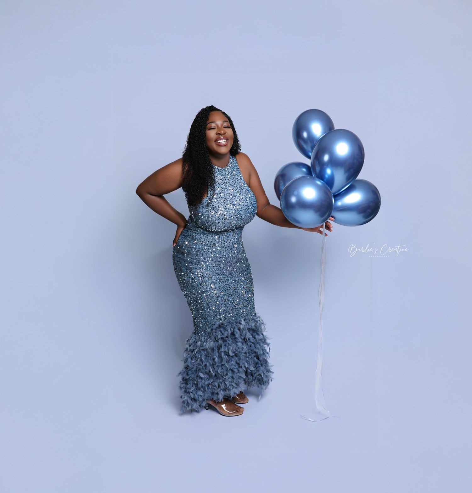 Smiling woman in blue sequin feather gown holds chrome blue balloons in studio.