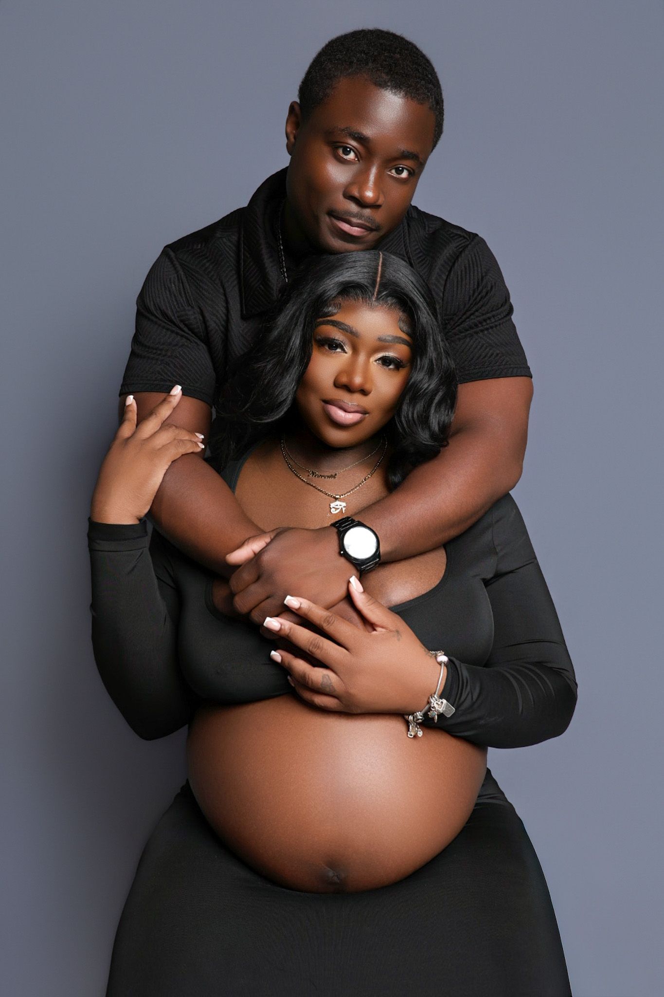 Couple maternity portrait in matching black outfits with man embracing pregnant woman from behind.