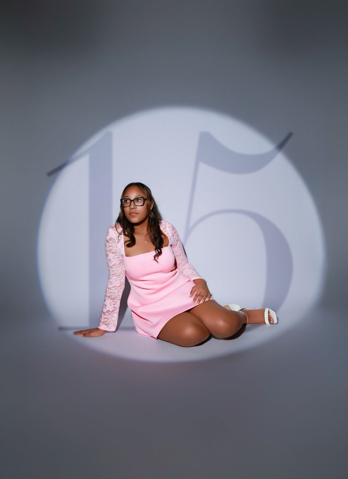 Young woman in pink lace dress sits in spotlight with number 15 projected behind her.