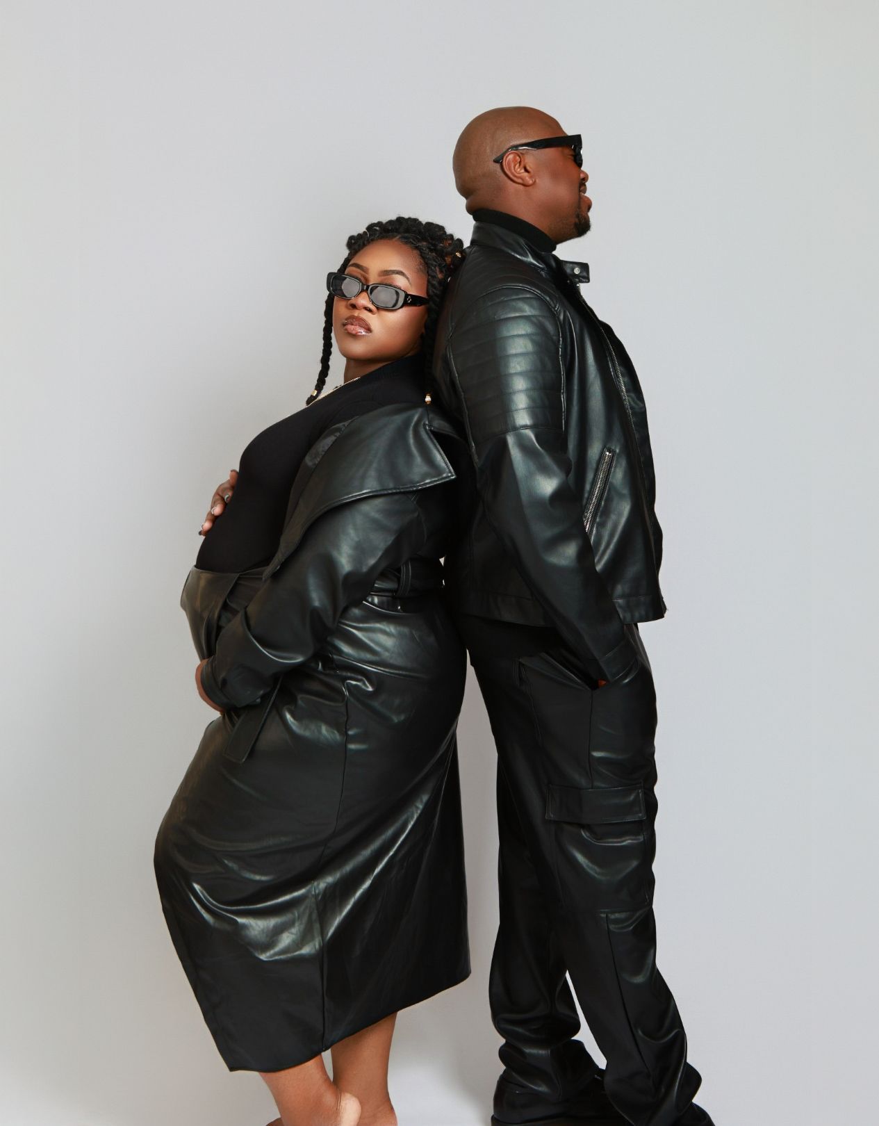 Man and woman in black leather outfits and sunglasses strike a back-to-back pose in a studio setting.