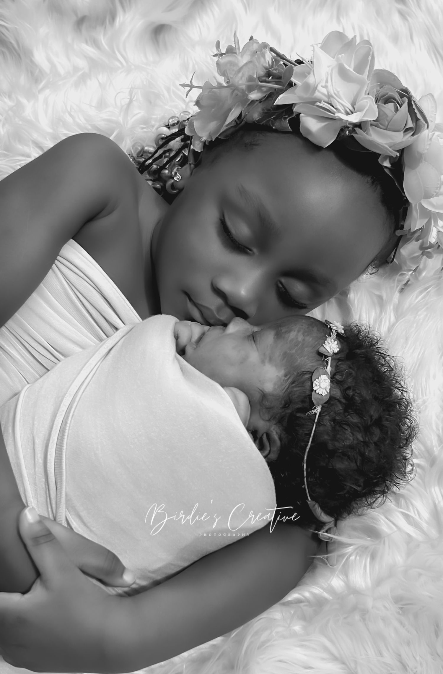 Black and white photo of a young girl kissing a swaddled newborn baby sister.
