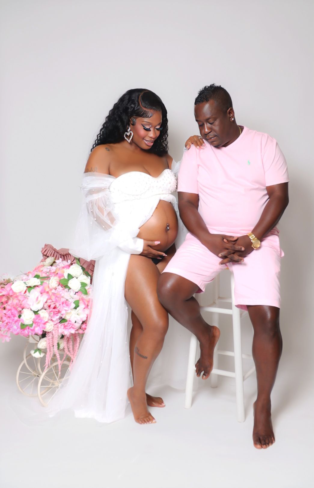 Maternity photo of couple in white and pink outfits with floral arrangement on white background.