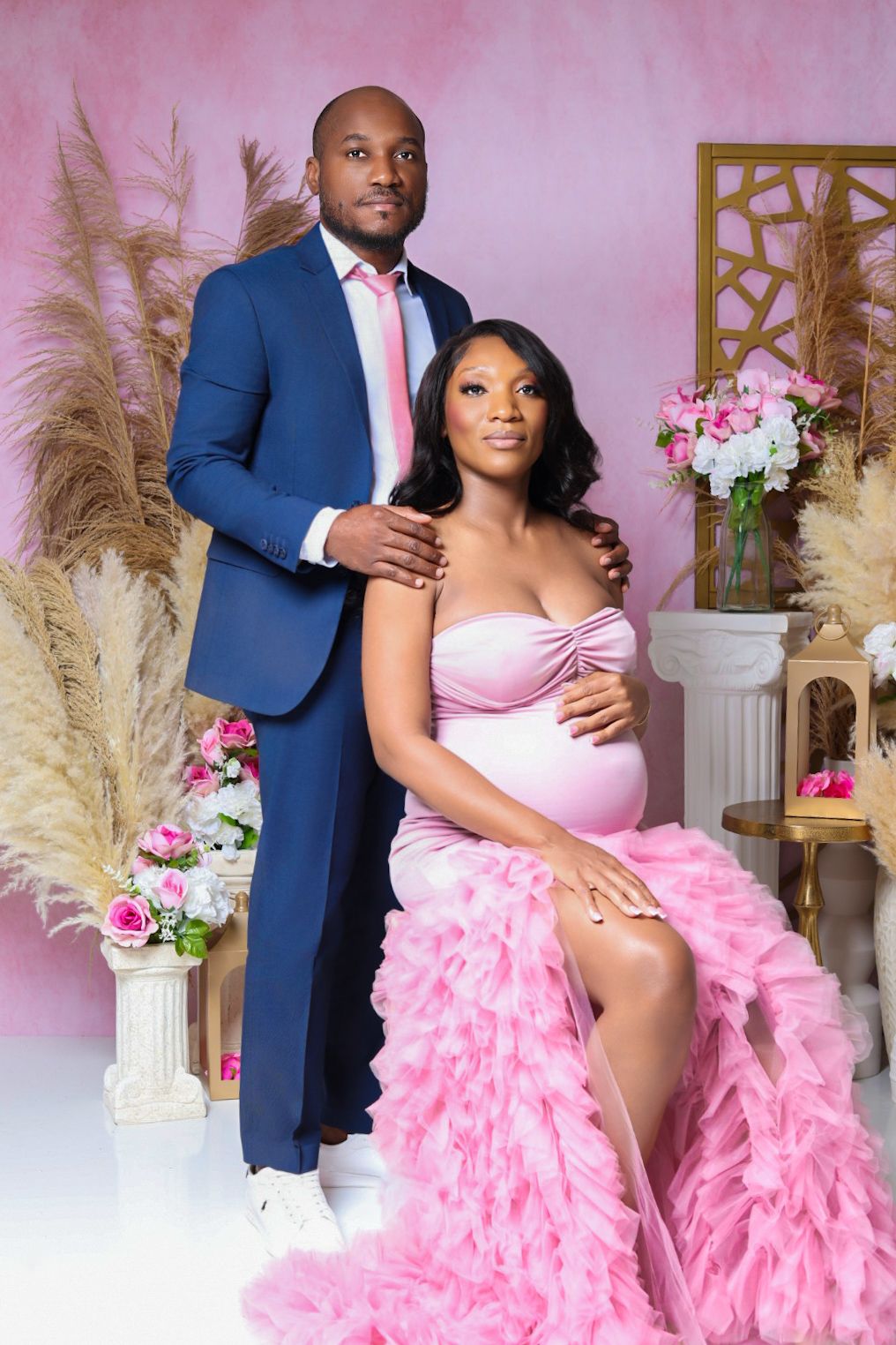 Elegant maternity shoot with woman in pink ruffled gown and man in navy blue suit on pink backdrop.