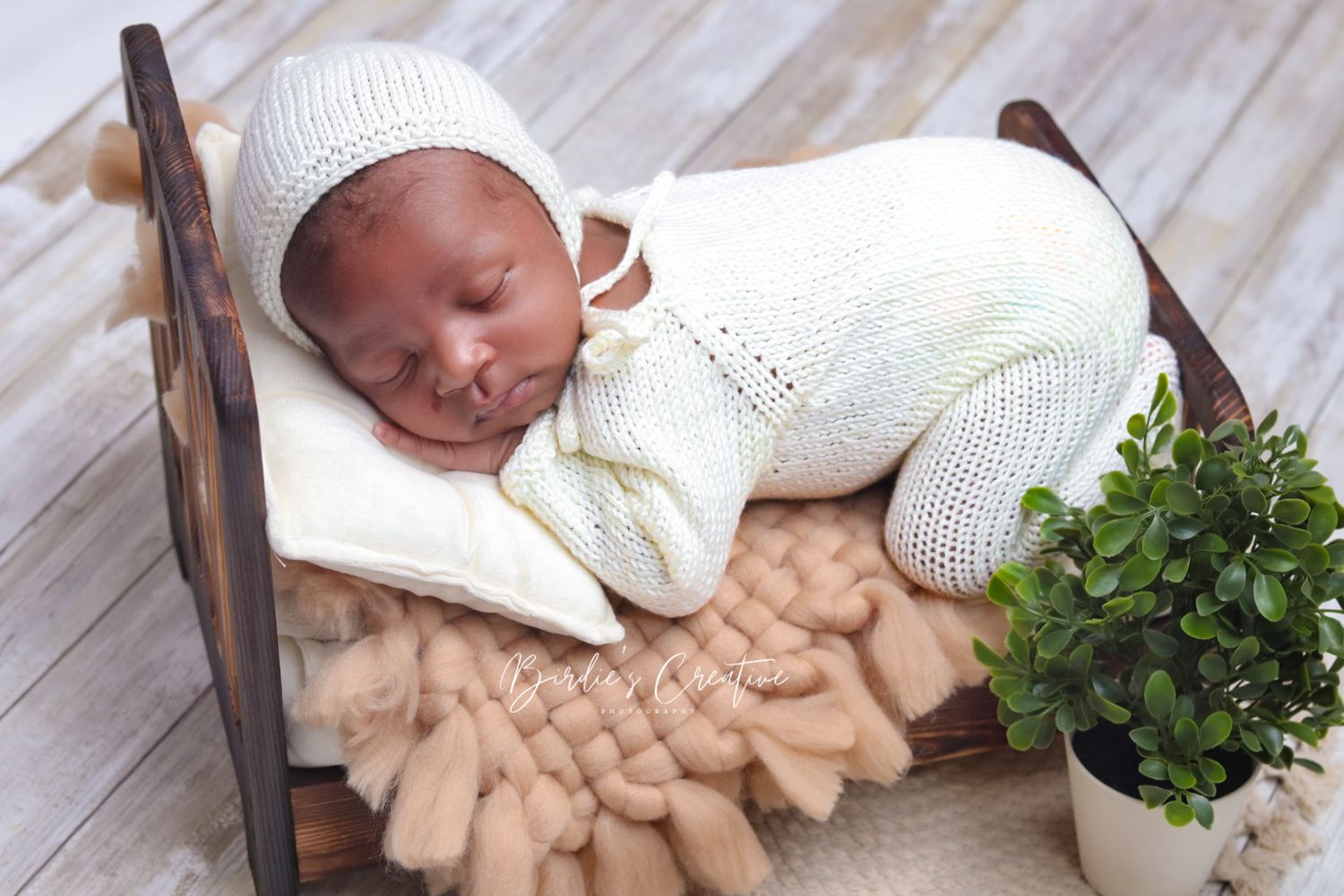 Sleeping newborn in cream knit outfit and bonnet rests in wooden bed with green plant decor.