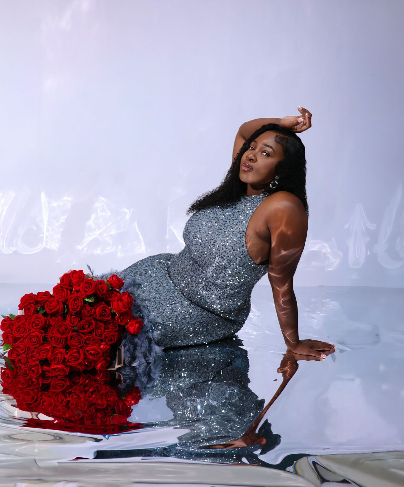 Woman in silver sequin gown poses dramatically with red roses on reflective studio floor.