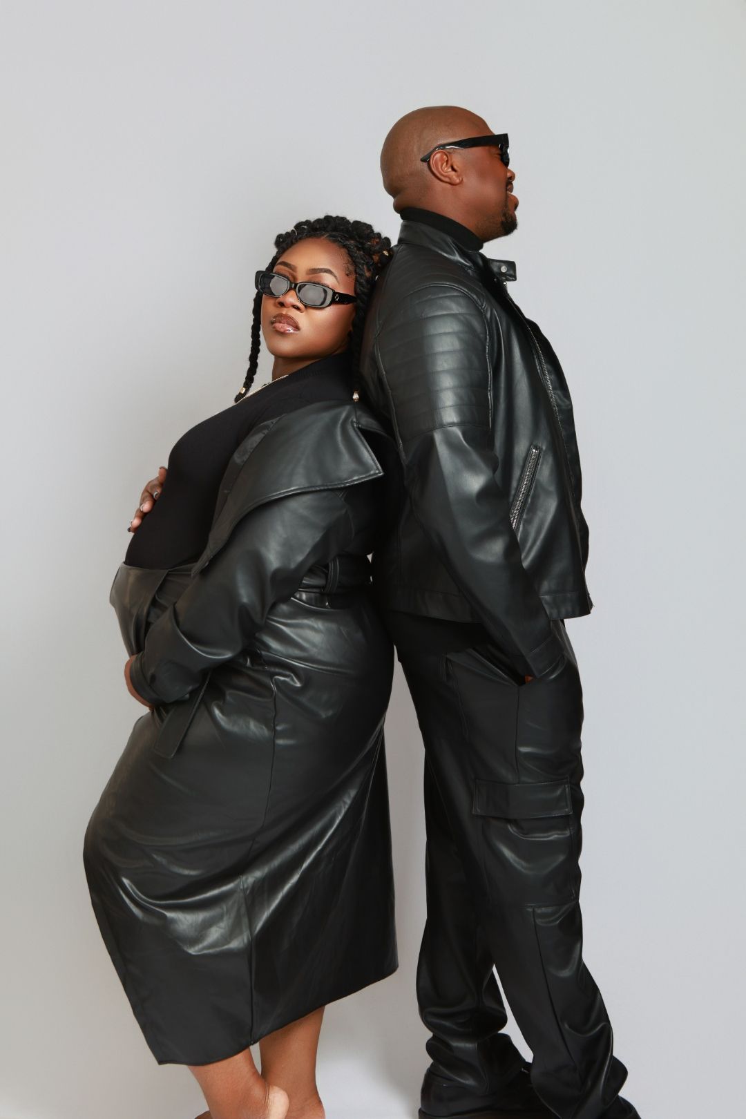Stylish couple in black leather coats and sunglasses pose back-to-back in a professional studio photo shoot.