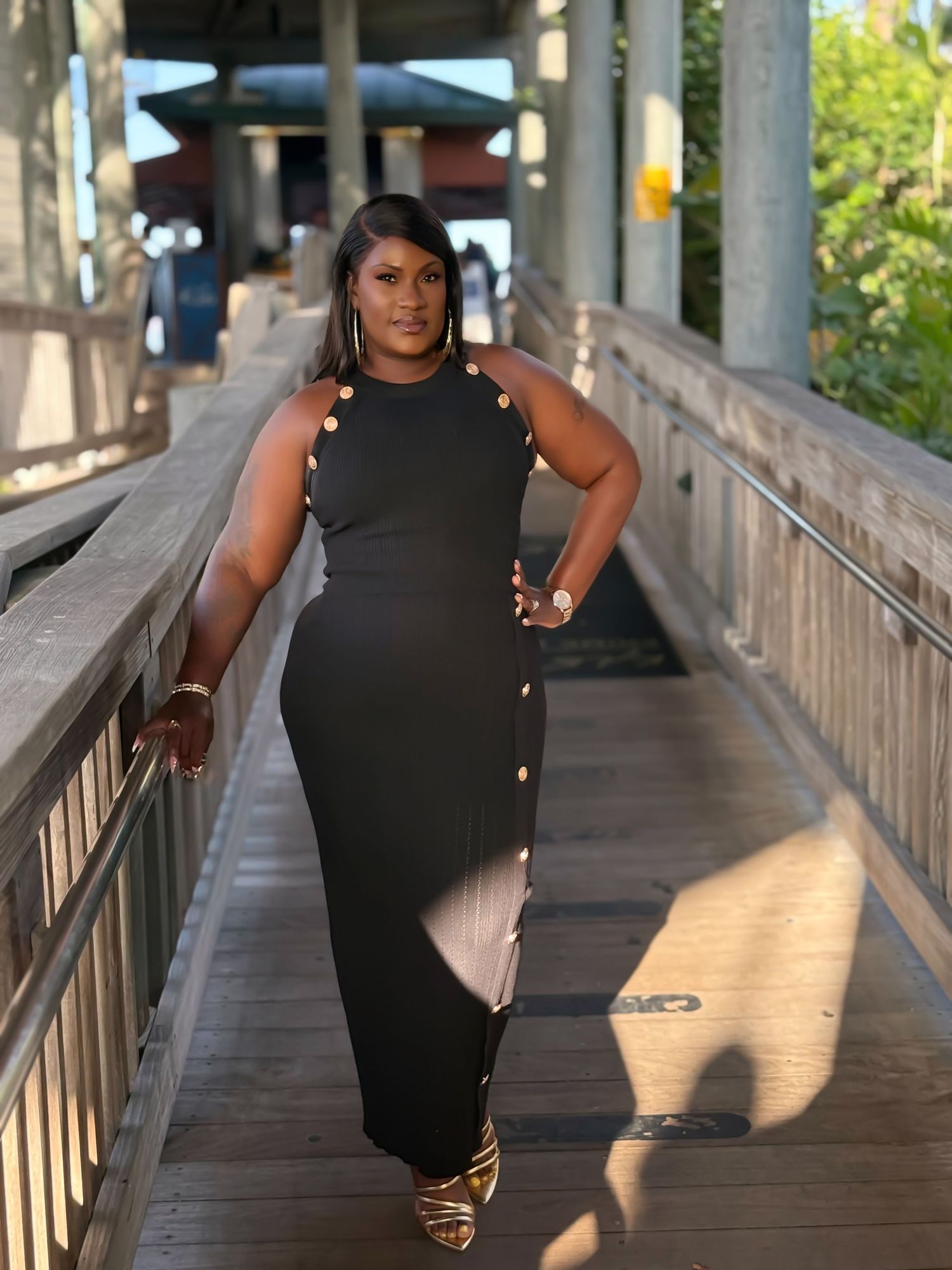Woman in black sleeveless maxi dress with gold buttons walks on wooden bridge outdoors.