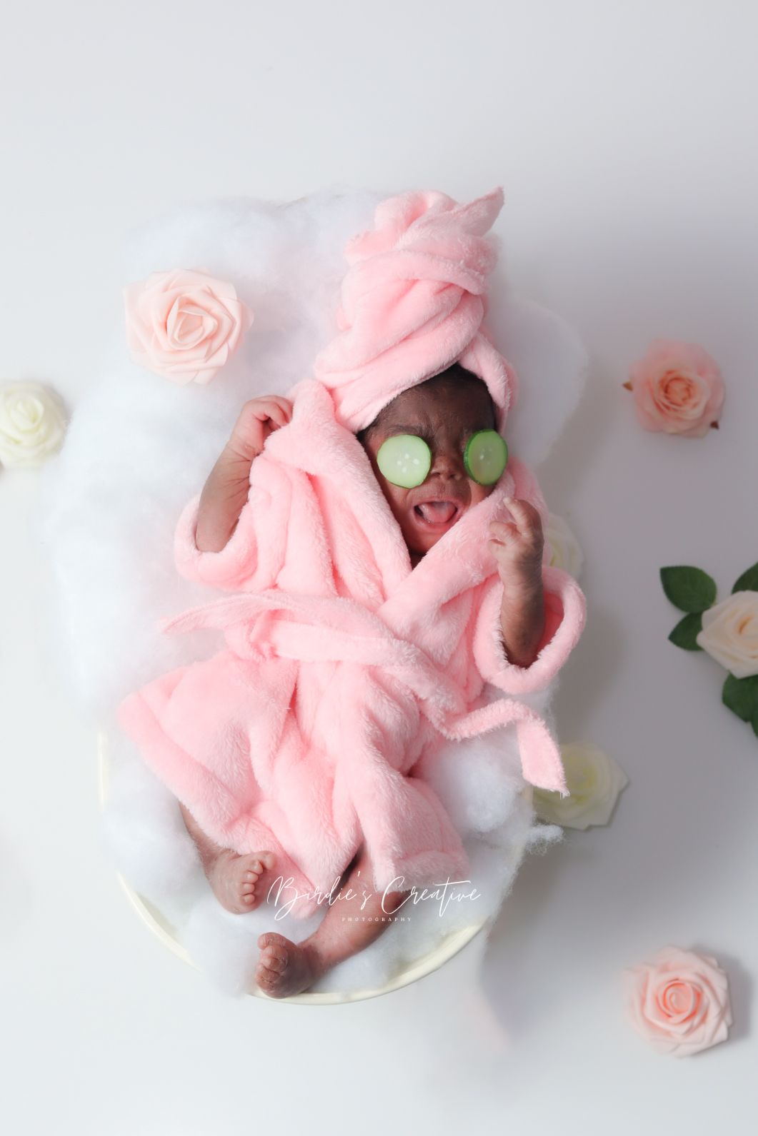Adorable newborn dressed in pink spa robe with cucumber eye mask poses in bubble bath setup.